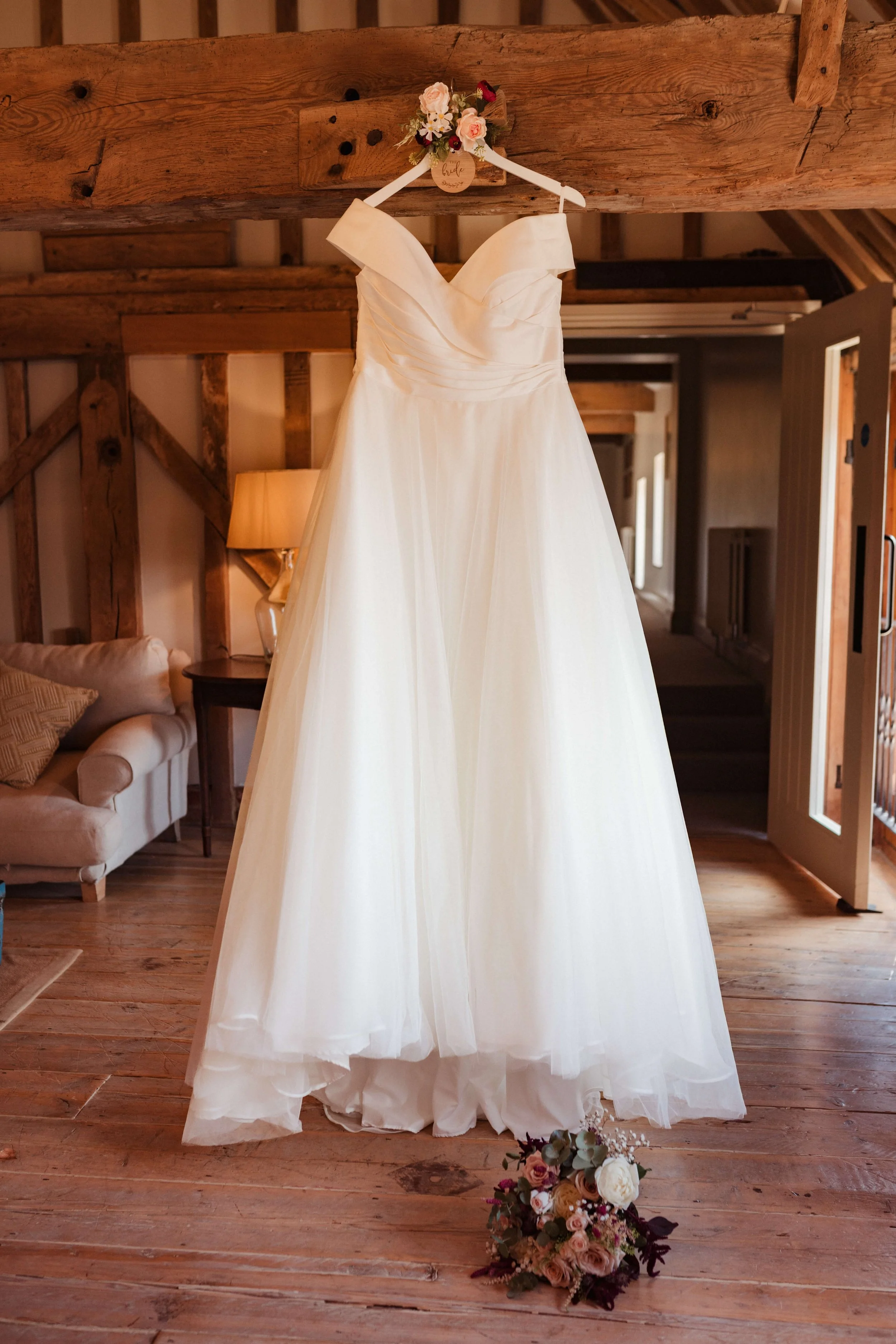 A white wedding dress hanging on a floral hanger in a rustic room with wooden floors and beams, with a bouquet of flowers on the floor.