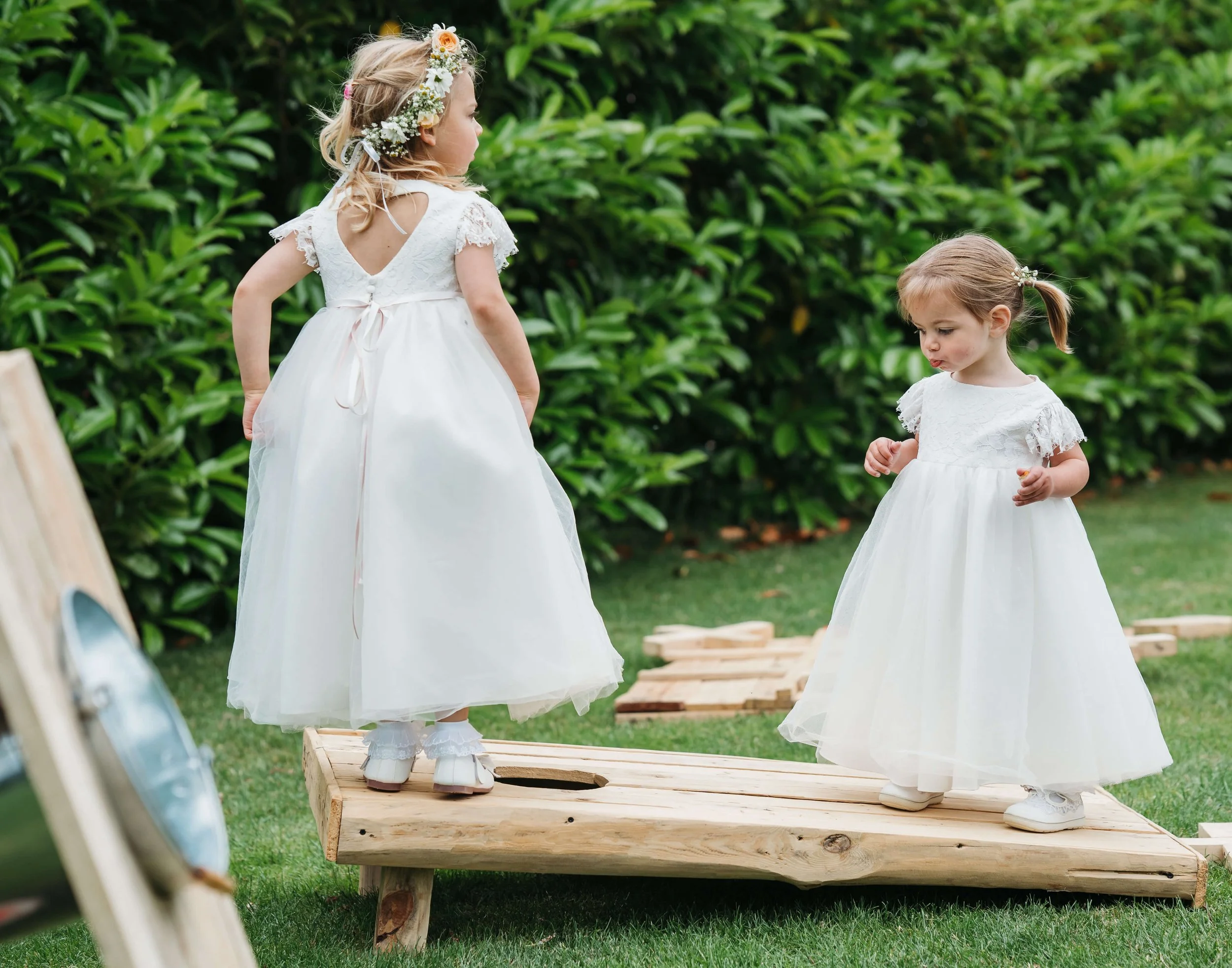 Two young girls in white dresses standing on a wooden platform outdoors, surrounded by greenery.