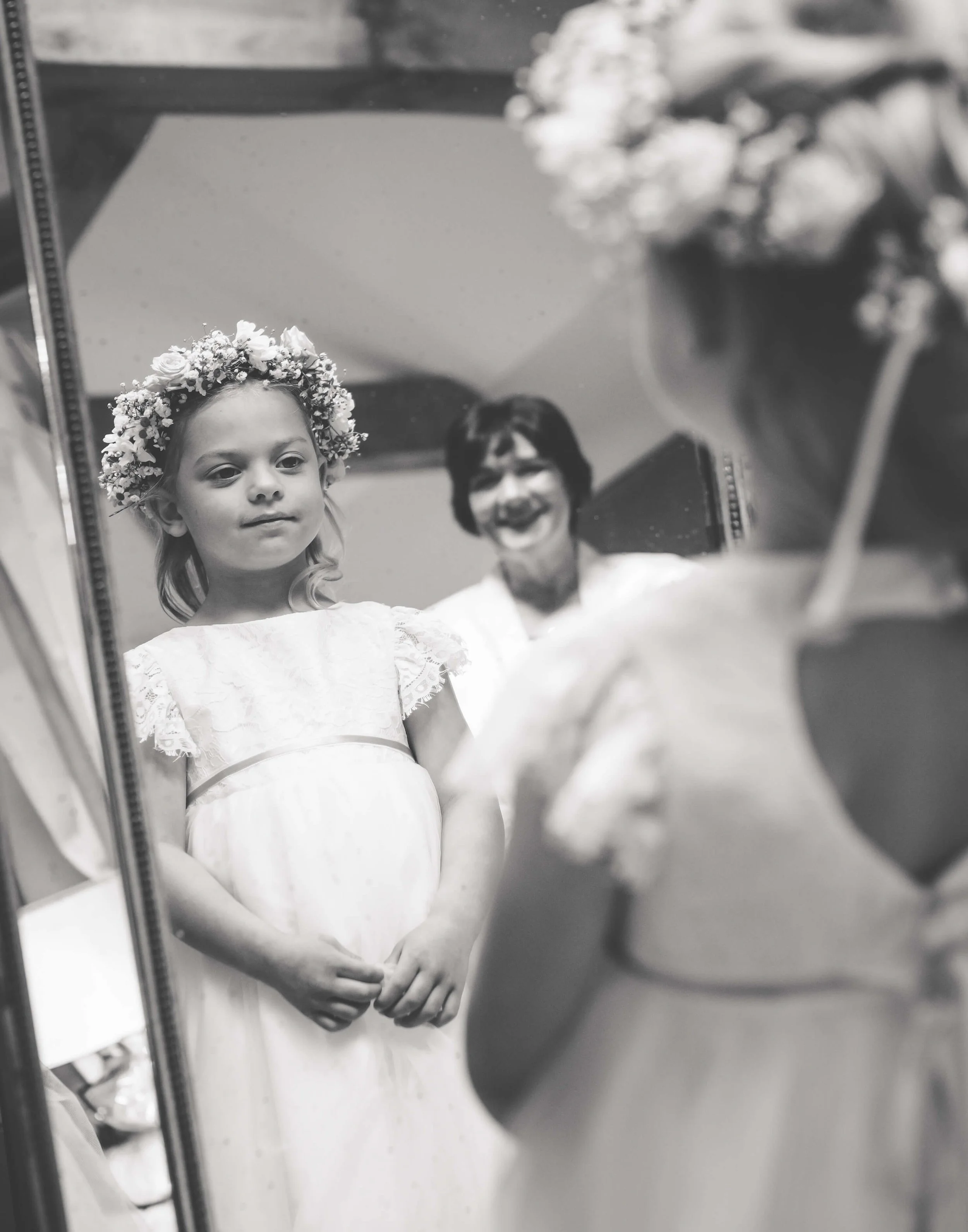A young girl in a white dress with lace details and a floral crown looking at her reflection in a mirror, with a woman smiling in the background.