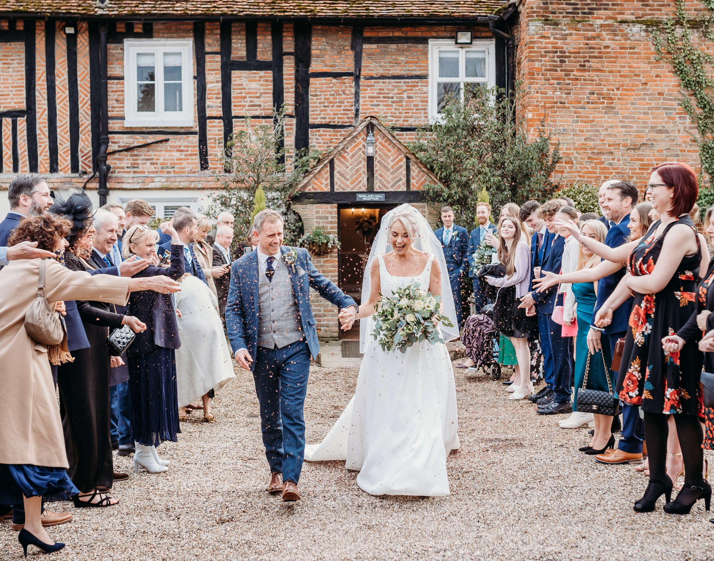 A bride and groom holding hands walking through a crowd of wedding guests outside a brick building, celebrating their wedding.