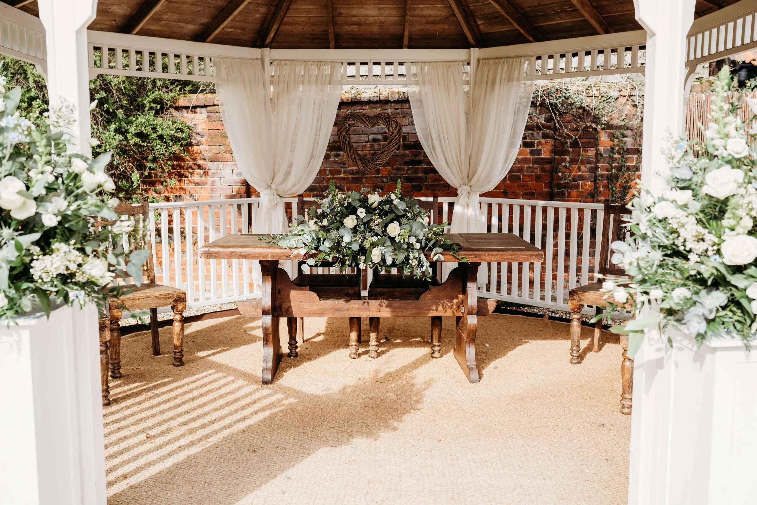 A wooden gazebo decorated for a wedding or special event, featuring white drapes, floral arrangements, a brick wall backdrop, and a heart-shaped decoration behind the table.