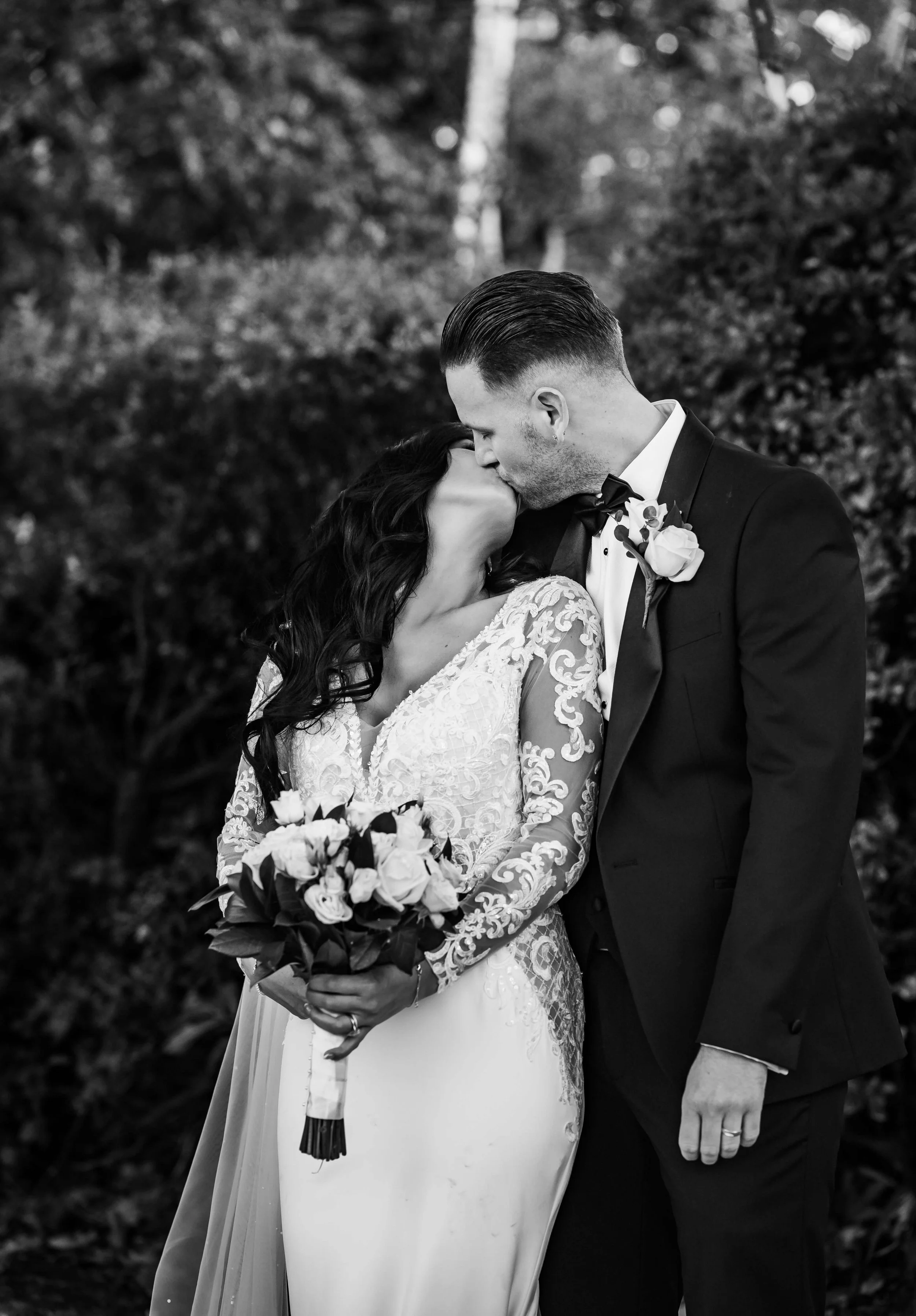 A bride and groom kissing during their wedding outdoors, the bride holding a bouquet of roses, both dressed in formal wedding attire, with a blurred natural background.