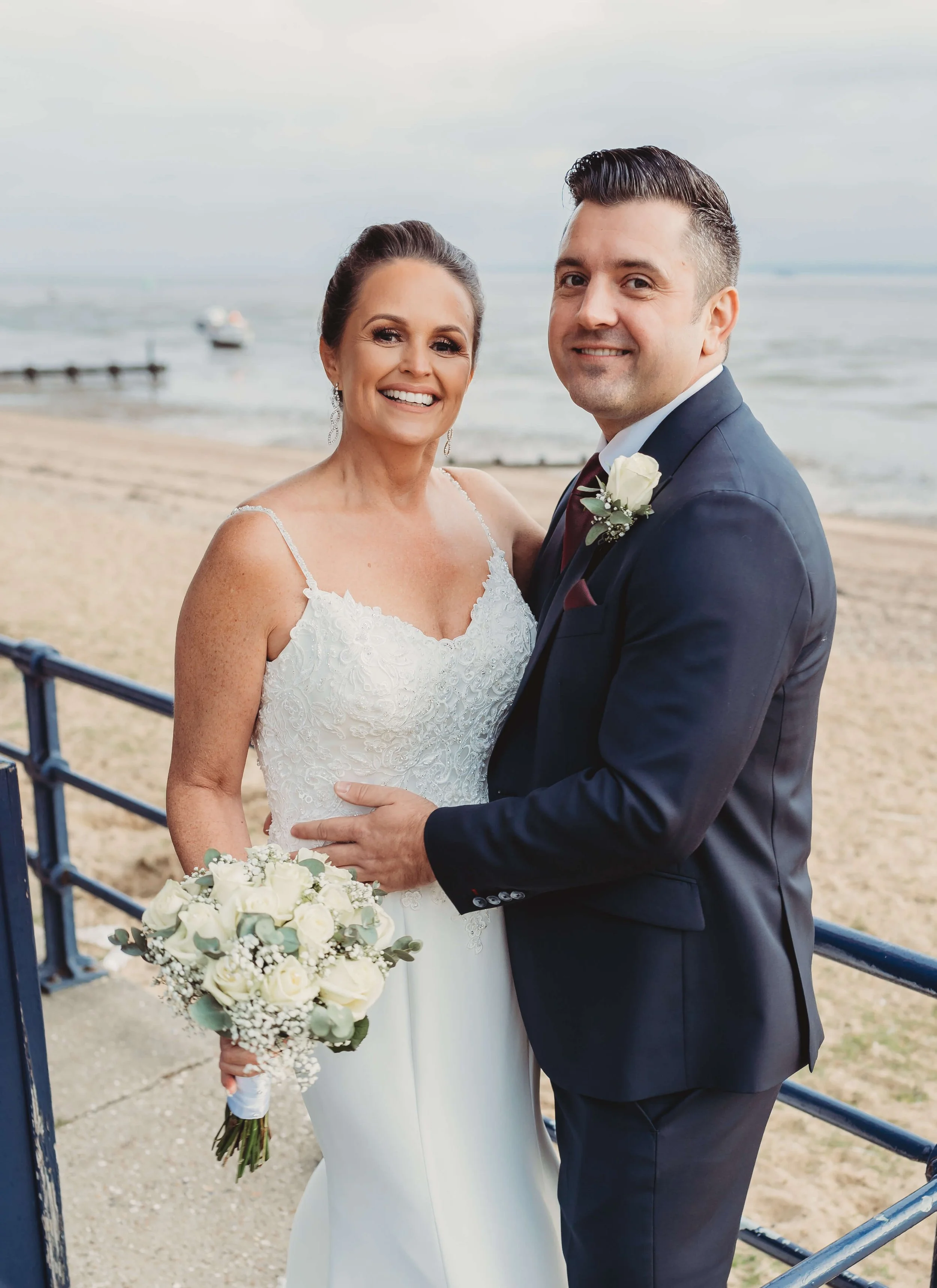A smiling bride in a white wedding dress with lace details and a groom in a navy suit, holding a bouquet of white roses, standing on a beach with the ocean in the background.