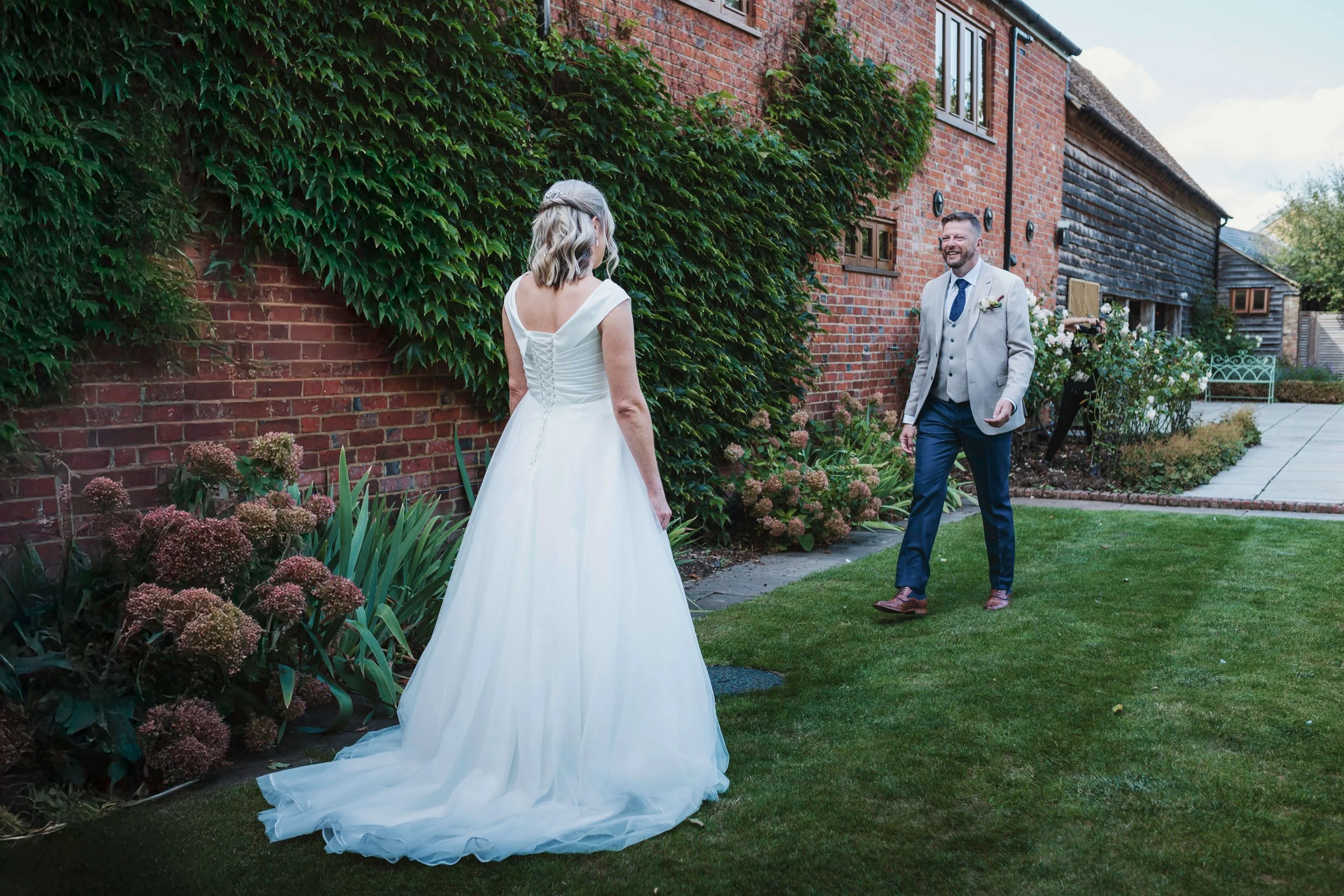 A bride wearing a white wedding gown faces a groom in a light gray blazer and green tie, both smiling, standing outdoors on a lush green lawn near a brick wall with green ivy and blooming plants.