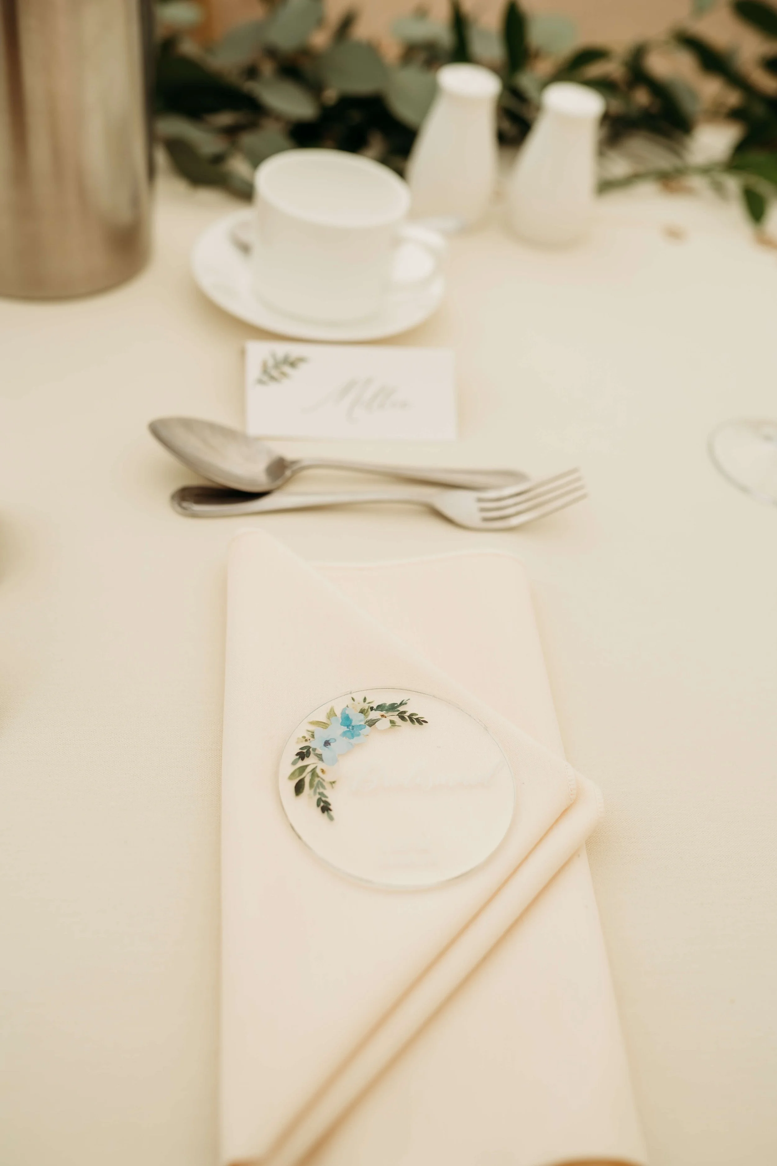 Elegant table setting with a white napkin, a floral name tag, and silverware on a cream-colored tablecloth.