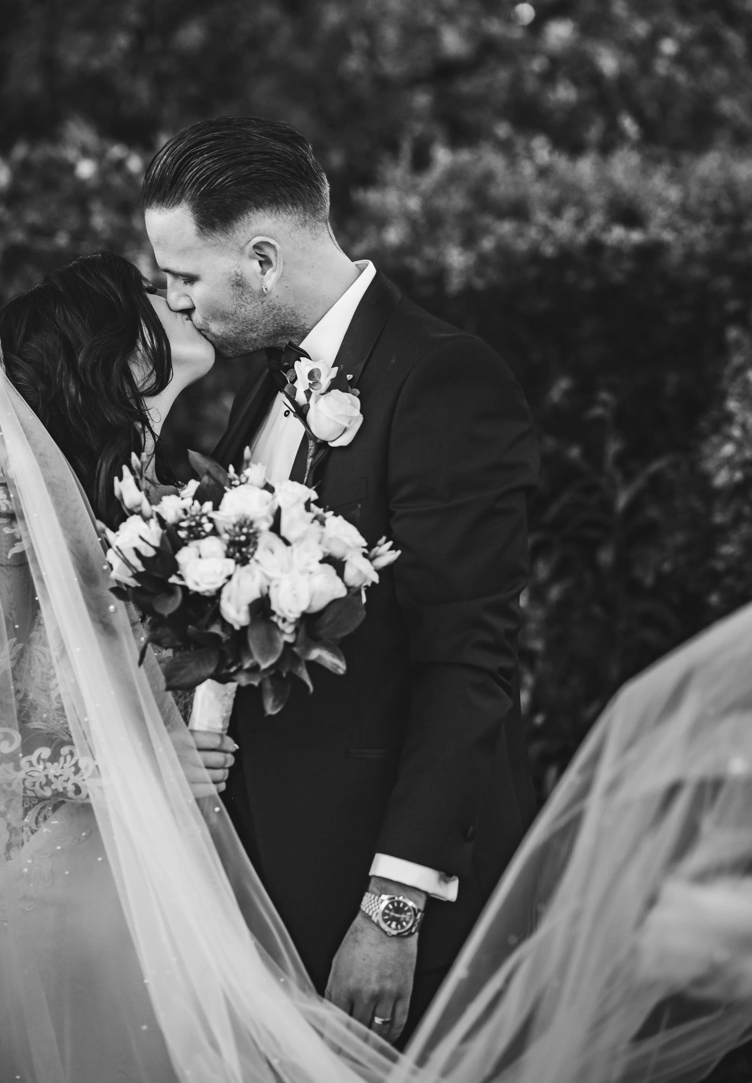 Black and white photo of a bride and groom sharing a kiss during their wedding ceremony, with the bride holding a bouquet of flowers.