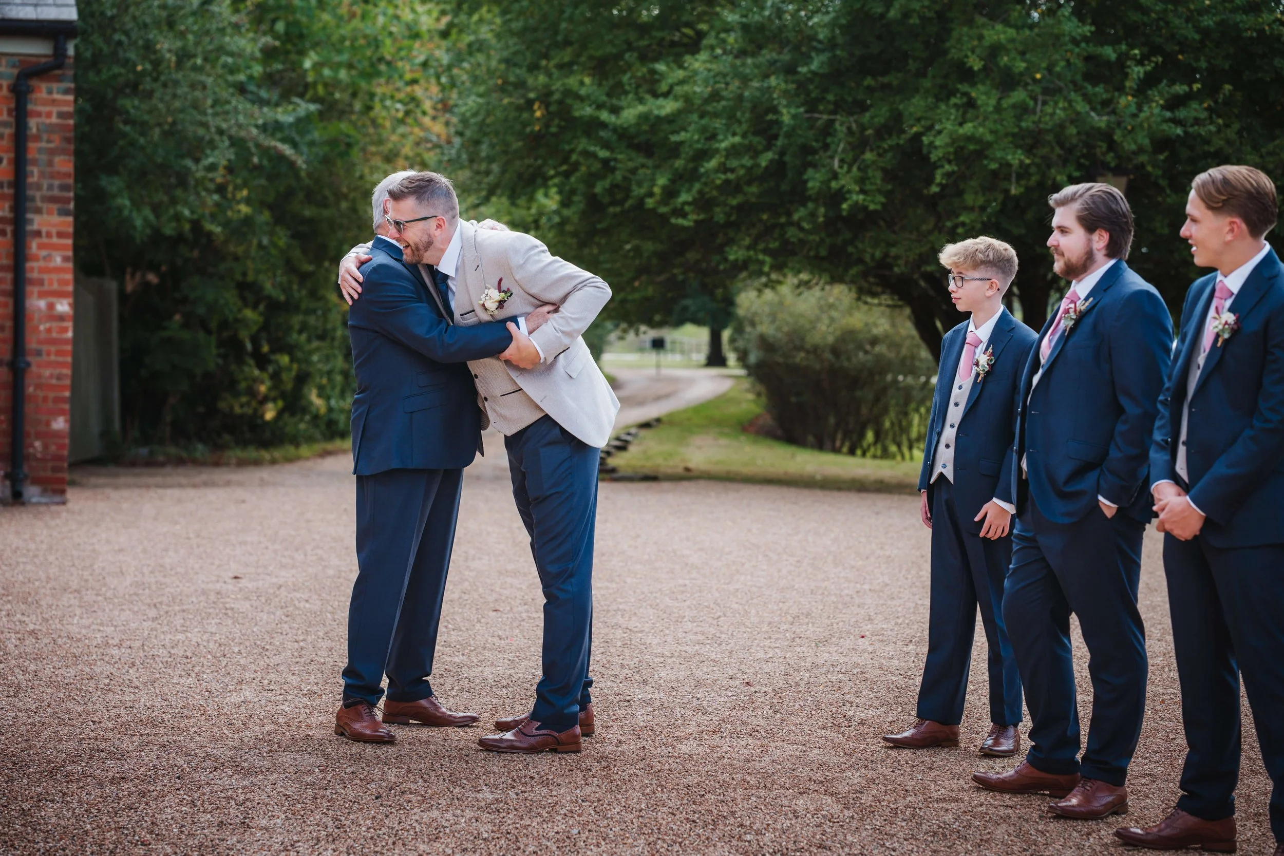 Two men hugging during a wedding ceremony outdoors, with three groomsmen dressed in blue suits standing nearby and smiling, on a gravel pathway with green trees in the background.