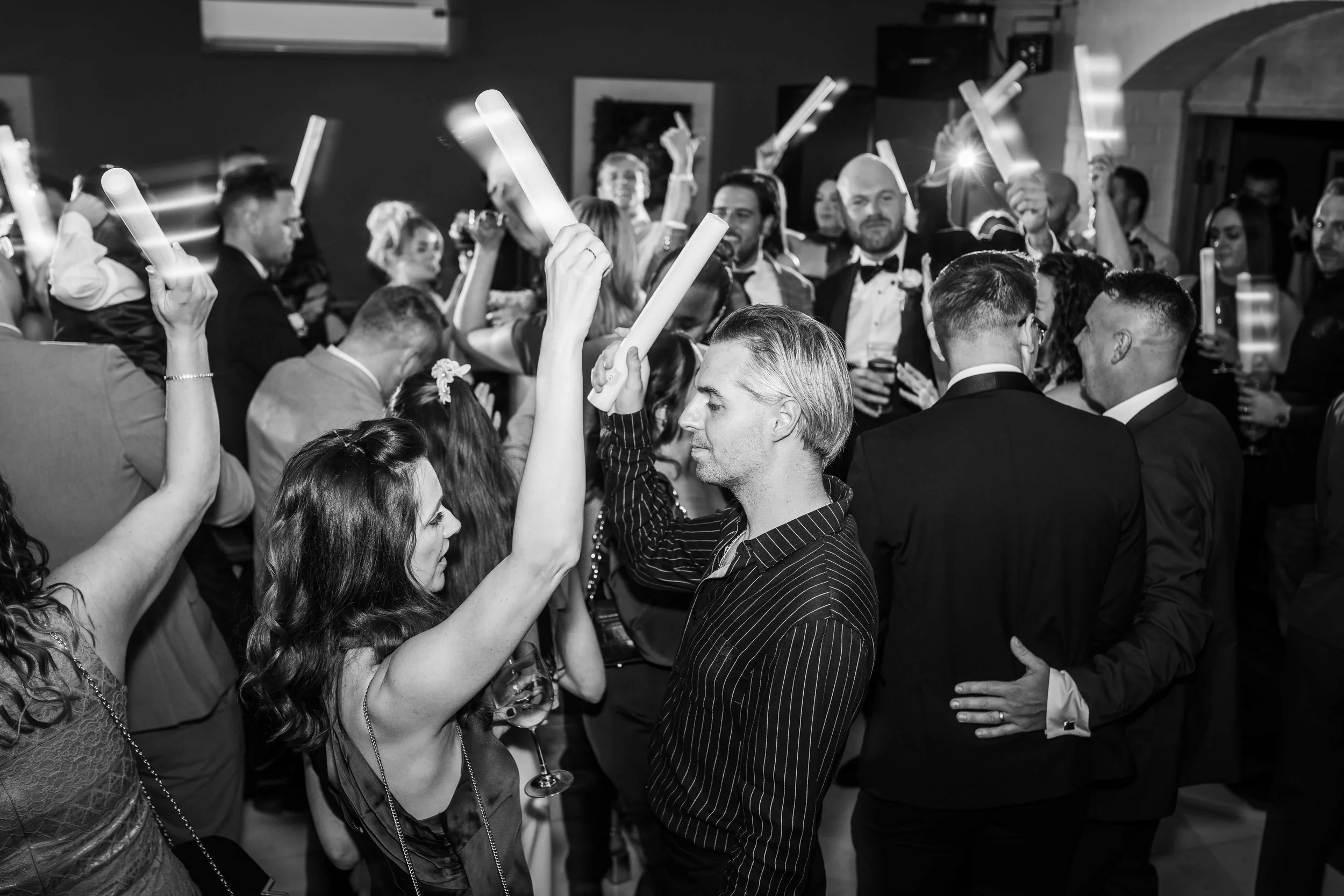 A black and white photo of people at a party or wedding reception dancing and holding glow sticks, with some people drinking wine and others talking closely.