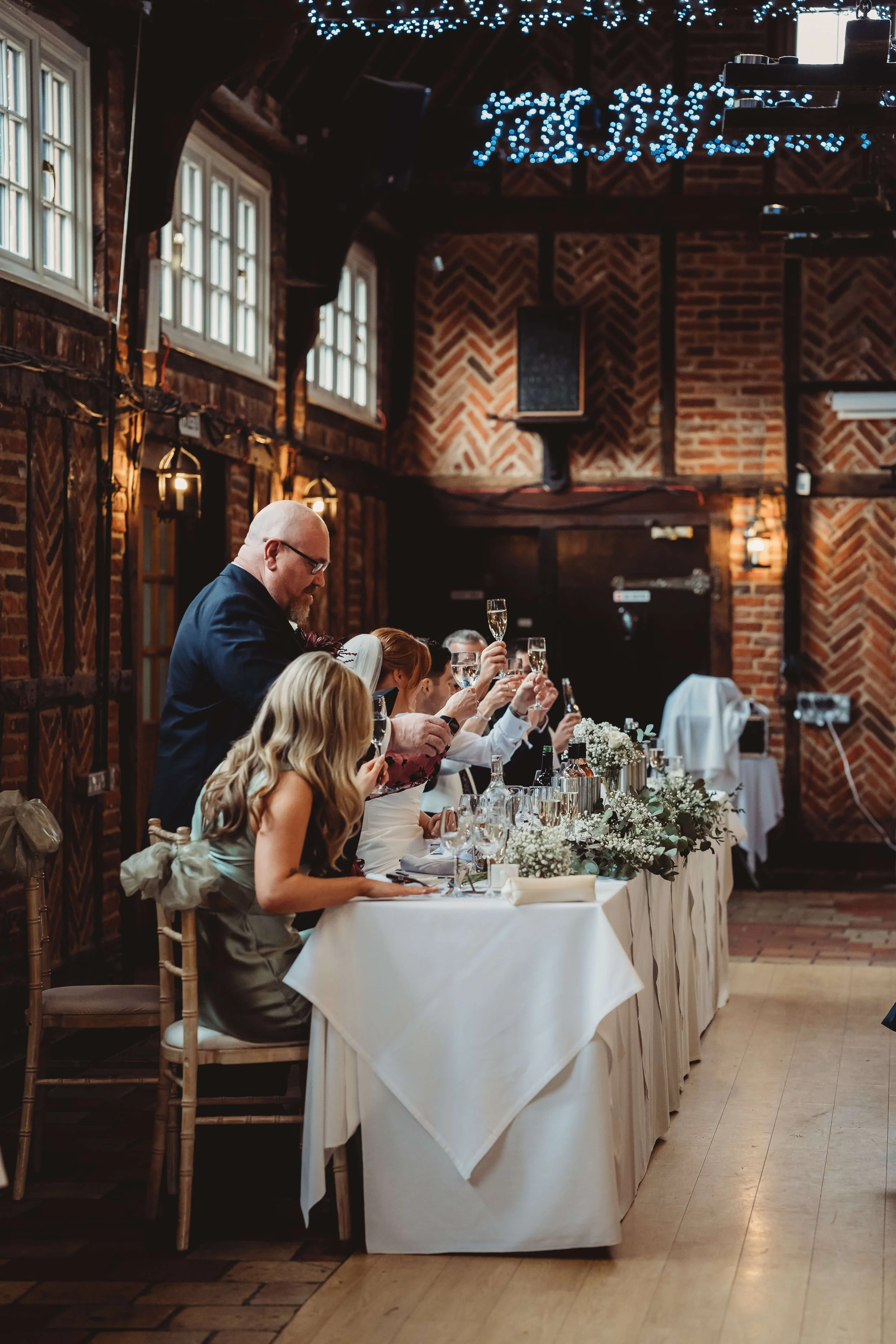 People at a wedding reception raising glasses in toast at a decorated banquet table in a rustic venue with exposed brick walls and string lights.