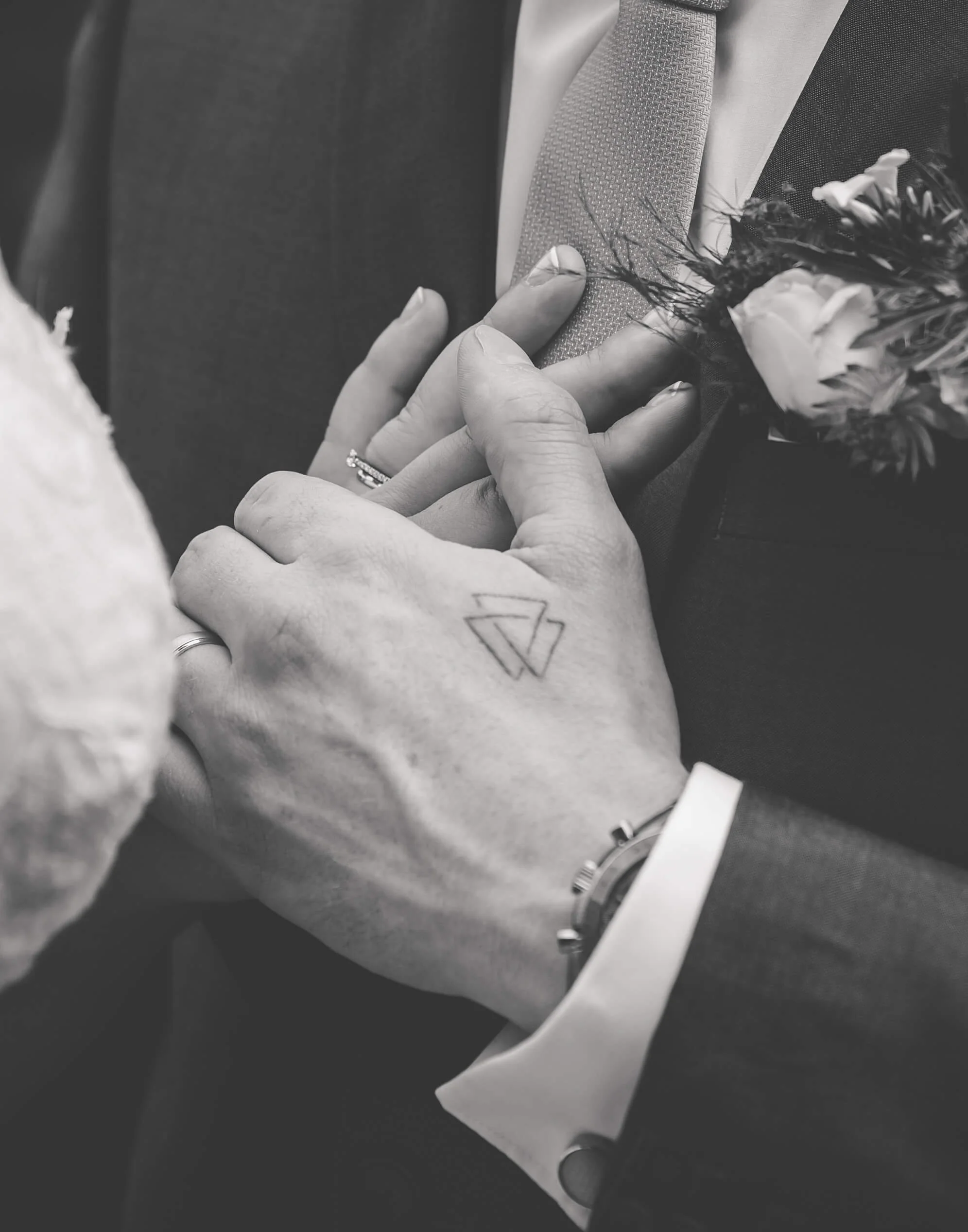 Close-up of a couple holding hands during a wedding, with wedding rings, a tattoo, and a boutonniere visible, in black and white.
