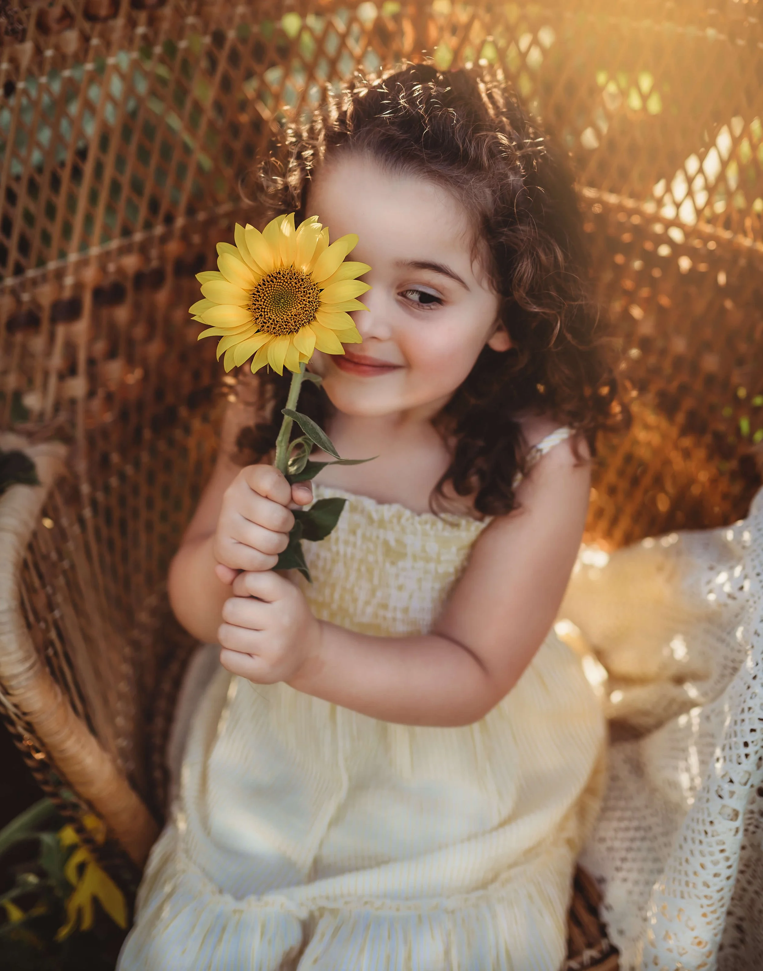 A young girl with curly hair in a yellow dress, sitting in a wicker chair, holding a sunflower near her face, smiling gently.