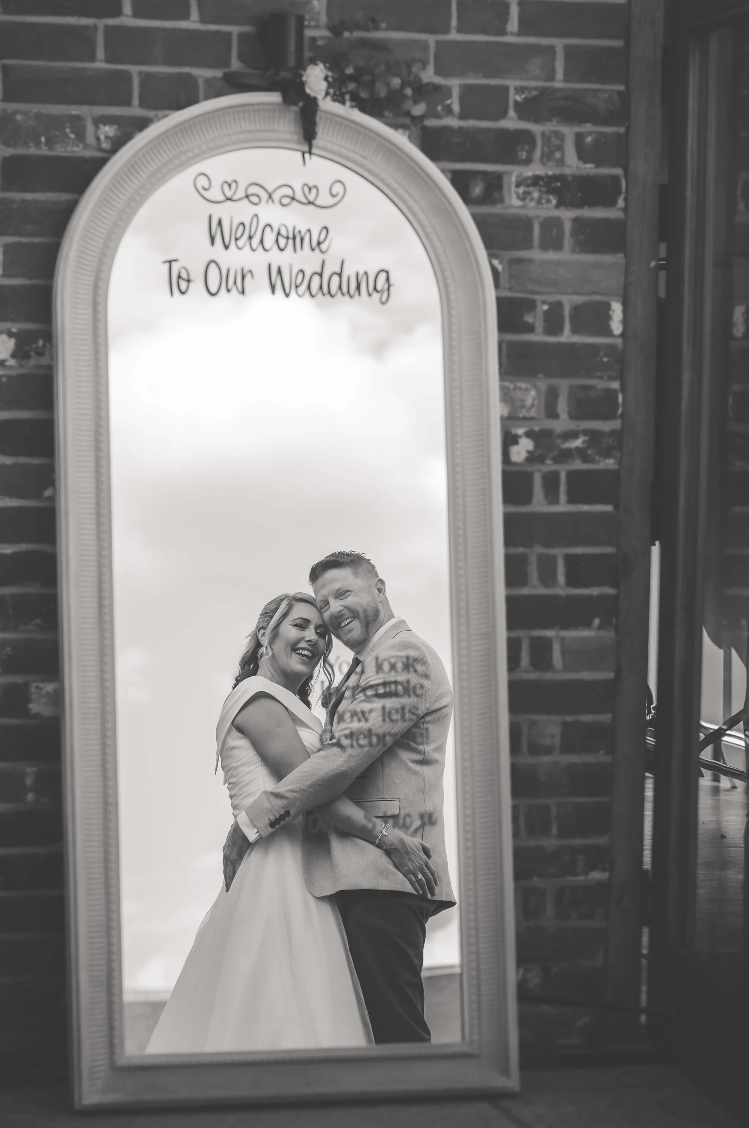 A black and white photo of a smiling bride and groom embracing in front of a large mirror with the text 'Welcome To Our Wedding' at the top. The mirror is placed against a brick wall.