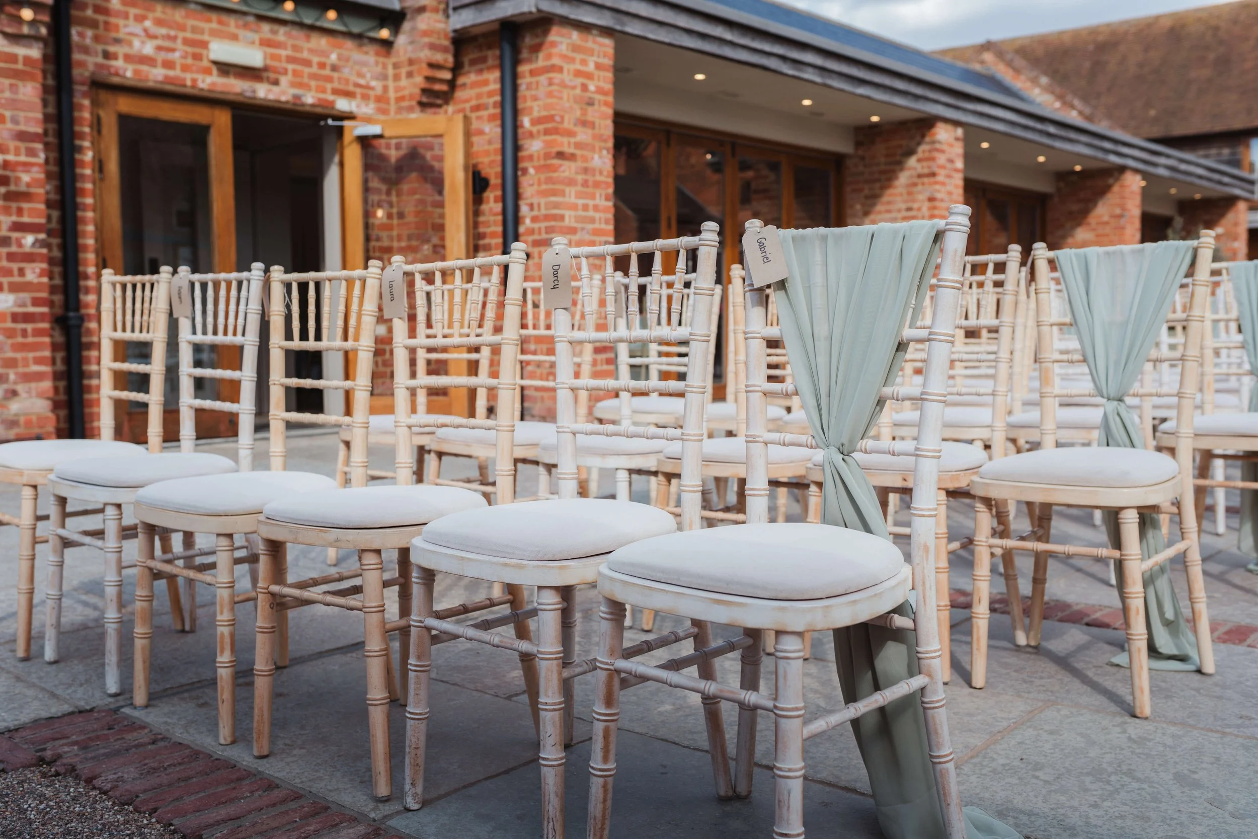 Rows of white chairs with pastel green fabric streamers tied to their backs, set up outdoors on a stone patio in front of a brick building for an event.