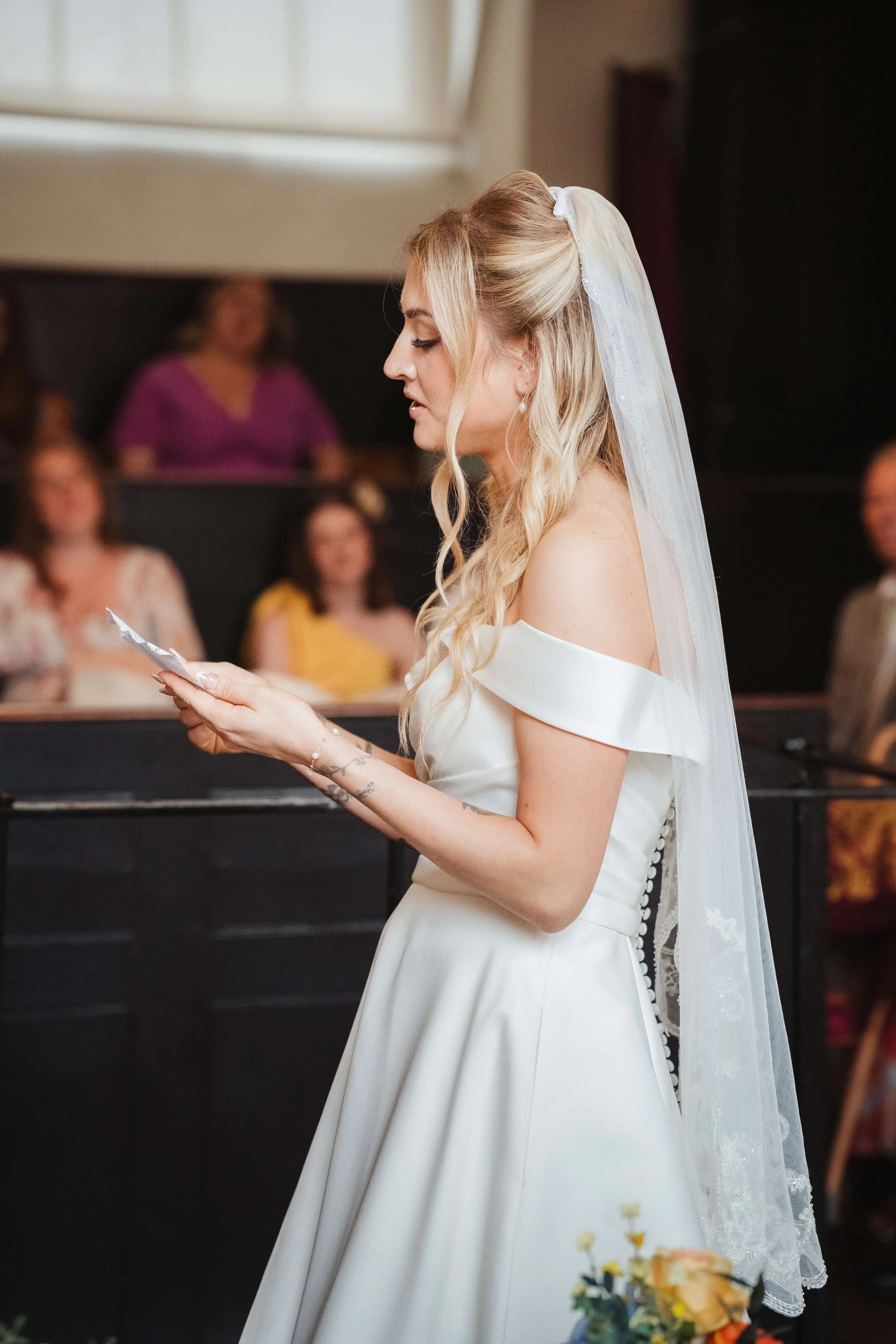 A bride in a white off-shoulder wedding dress and veil reading a note during her wedding ceremony.