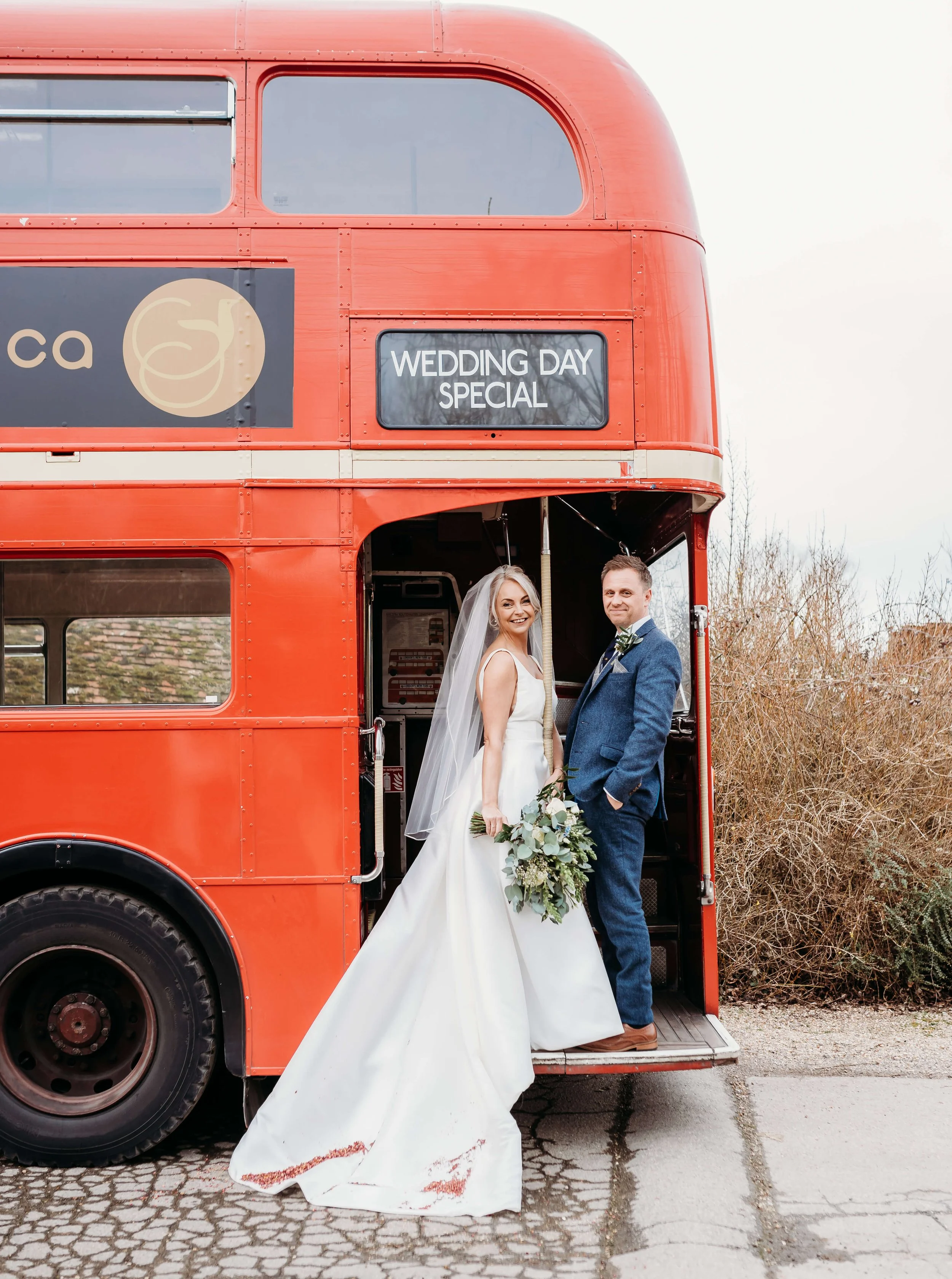 A bride and groom standing on the steps of a red double-decker bus, with a sign that says 'Wedding Day Special', outdoors with trees in the background.