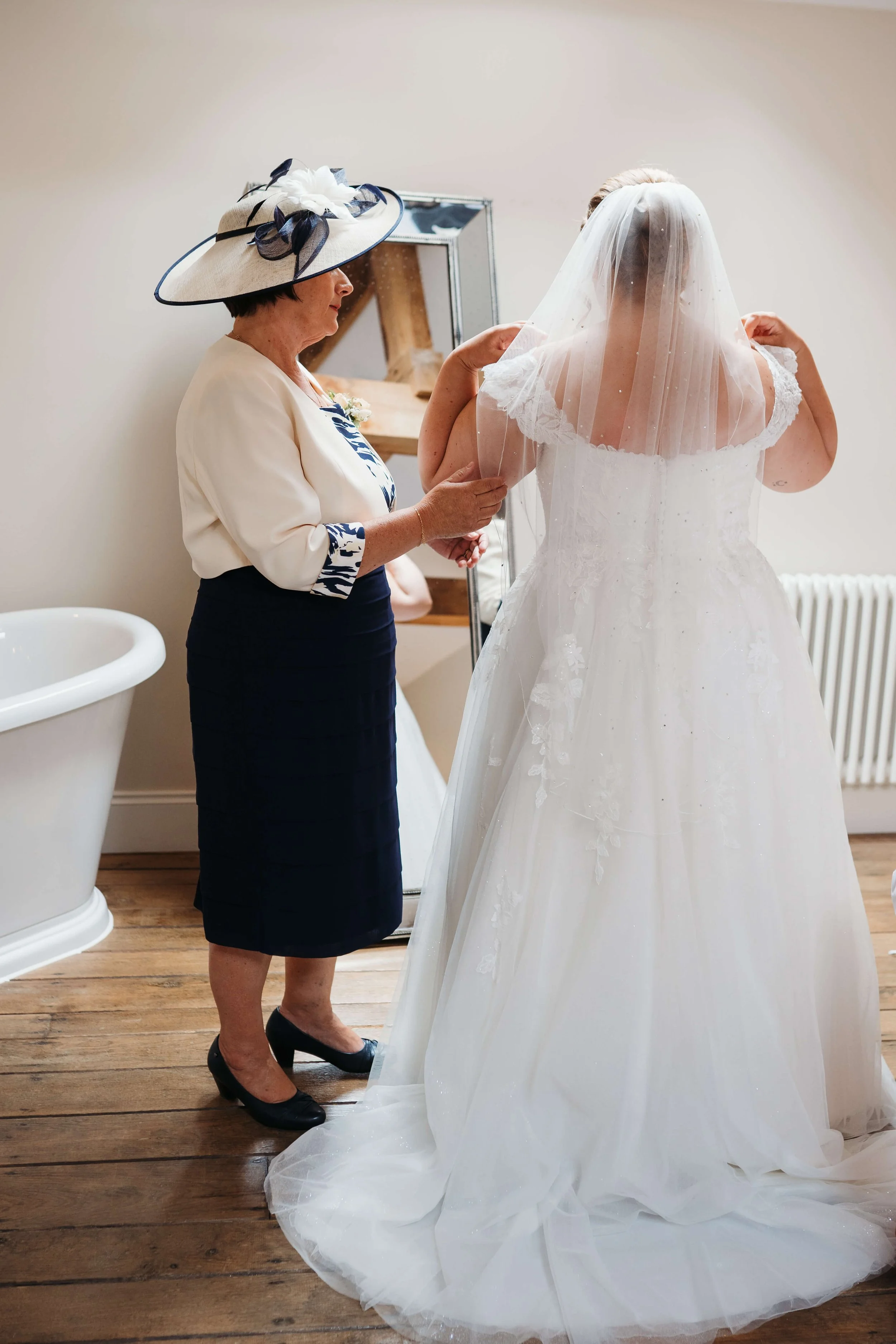 An elderly woman helping a bride adjust her wedding dress in a room with wooden floors and minimal decor.