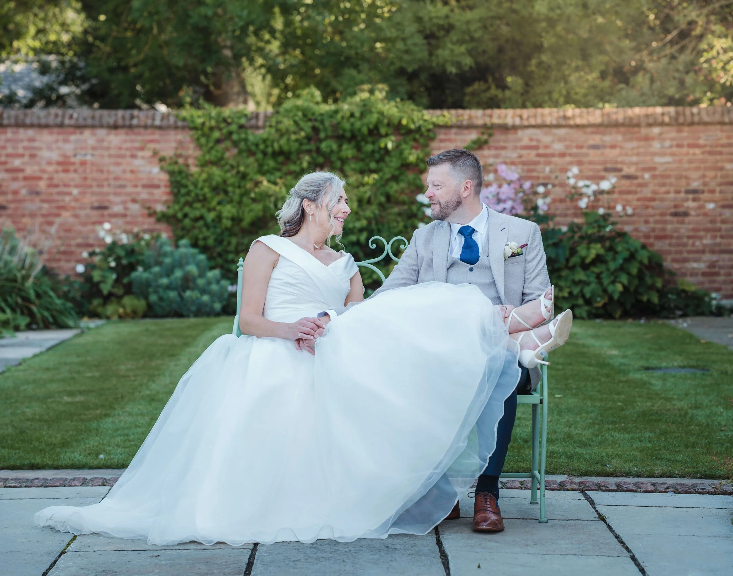 A bride and groom sitting on a green bench in a garden, smiling at each other, with the bride holding her wedding dress. The scene features a brick wall and greenery in the background.