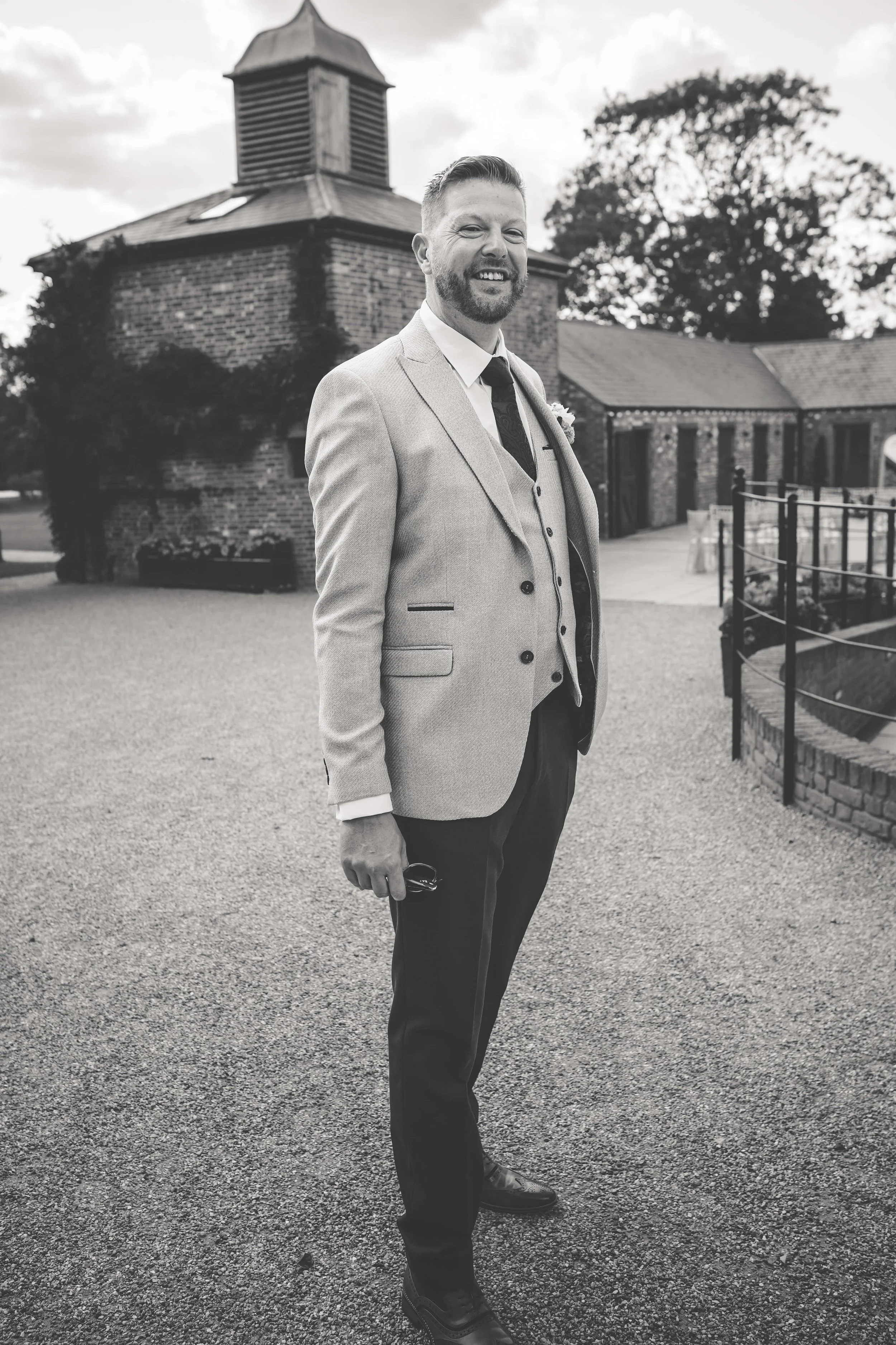 A smiling man in a light-colored suit jacket, dress shirt, and tie standing outdoors on a gravel path with a brick building and trees in the background.