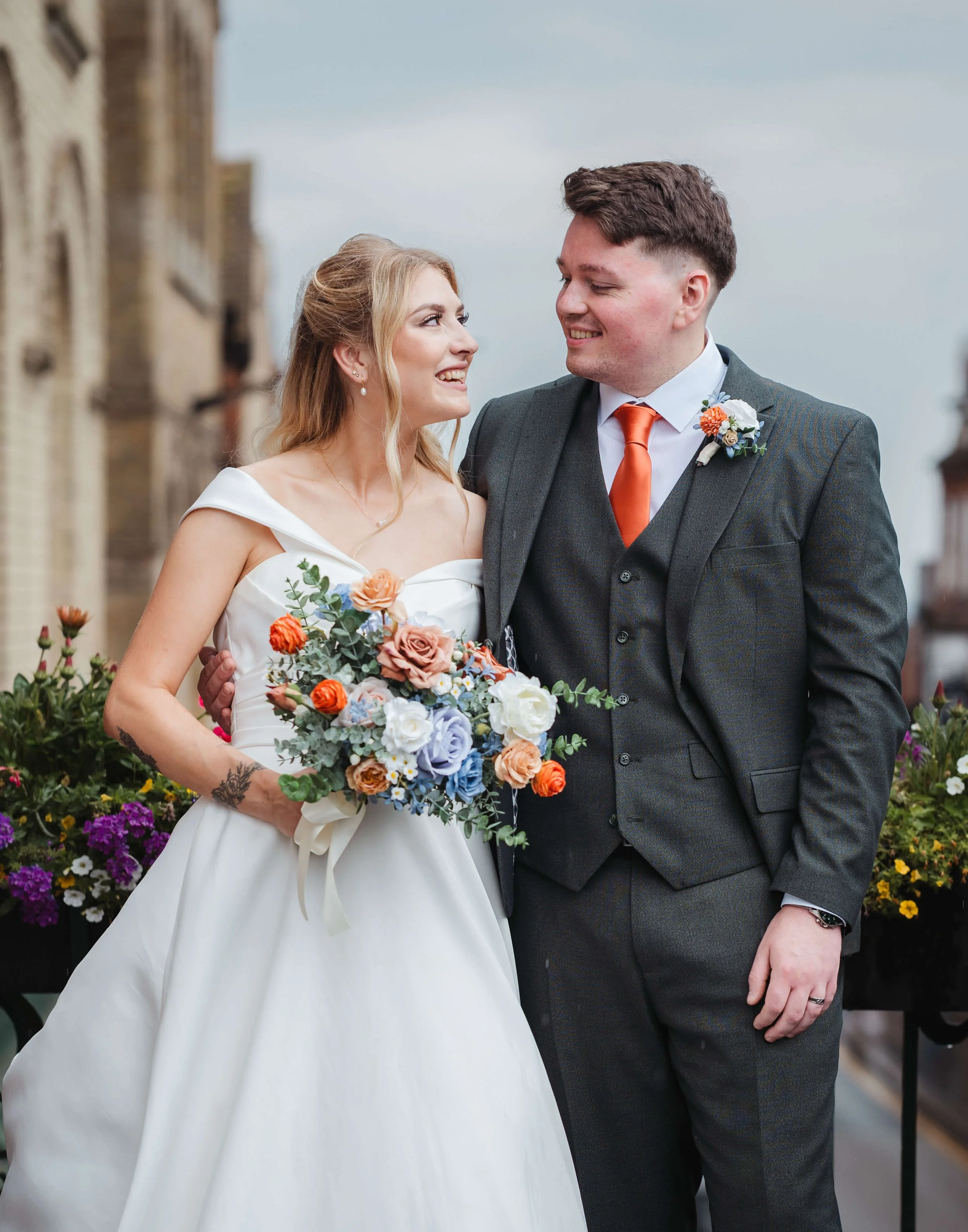 A bride and groom smiling at each other outdoors on their wedding day, with flowers and old buildings in the background.