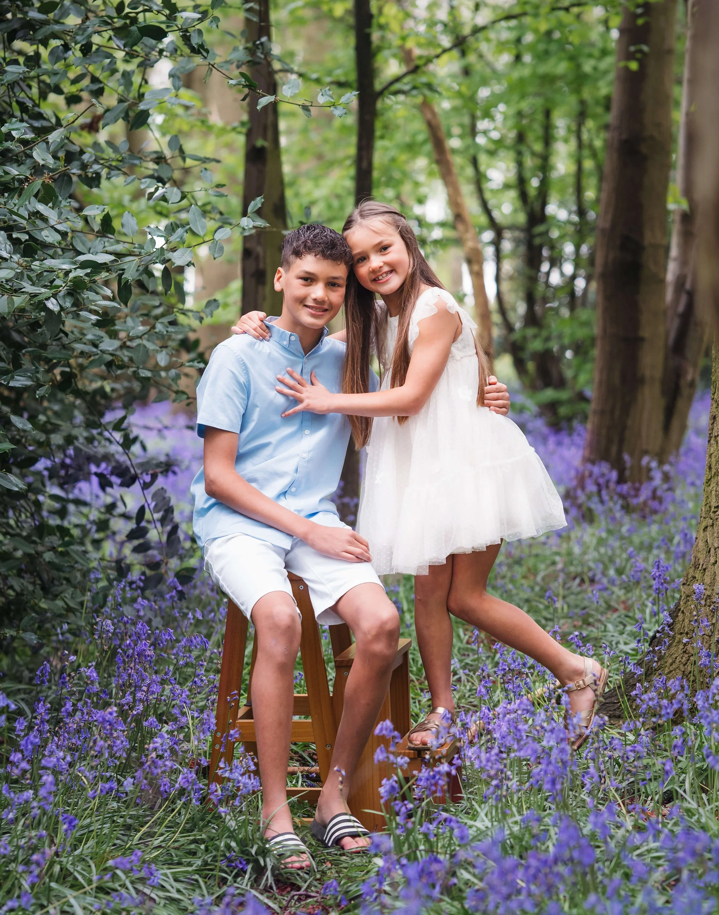 A boy and girl in a forest with purple flowers, smiling at the camera, the girl standing on a small platform. The boy is sitting on a wooden stool, wearing a light blue shirt and white shorts, while the girl is in a white dress.
