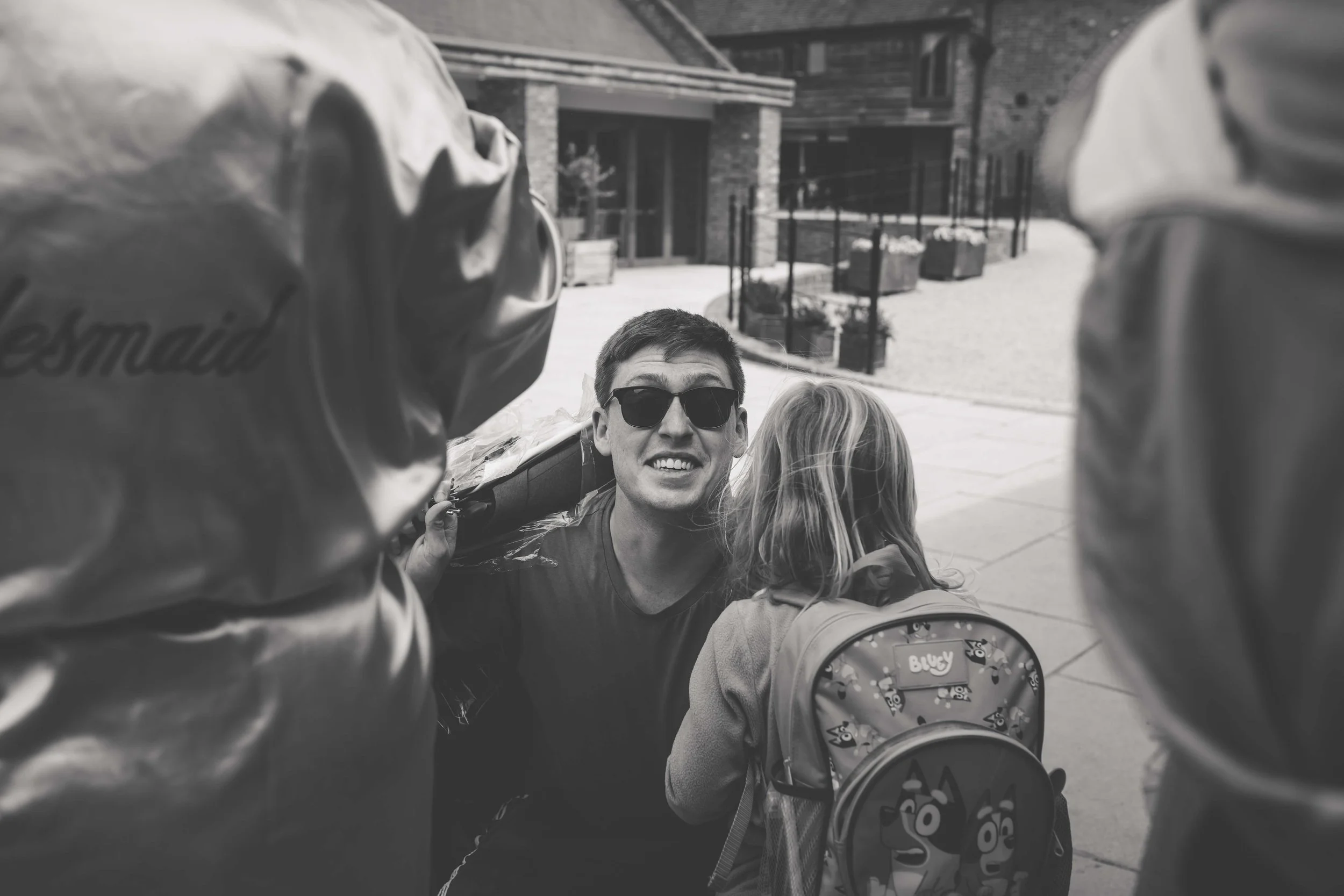 A young man wearing sunglasses and a casual shirt, smiling, is interacting with a young girl with long hair and a backpack, outdoors near a building.