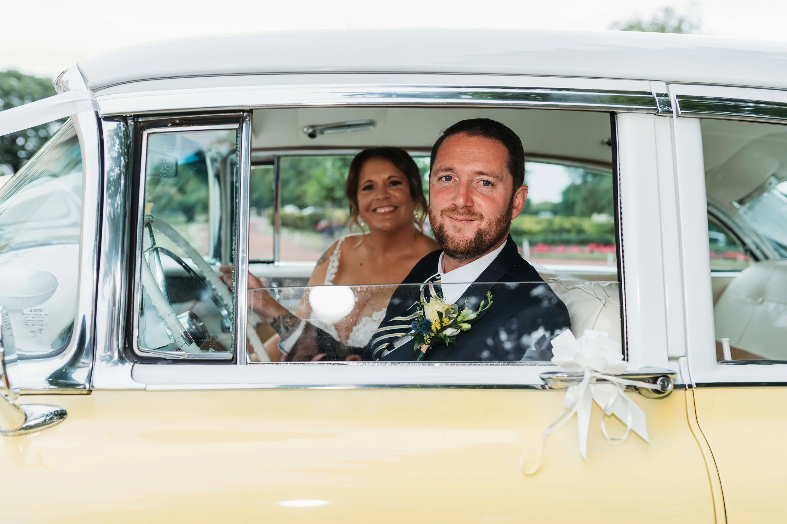 A bride and groom sitting inside a vintage yellow car decorated with white ribbons, smiling at the camera during a wedding celebration.
