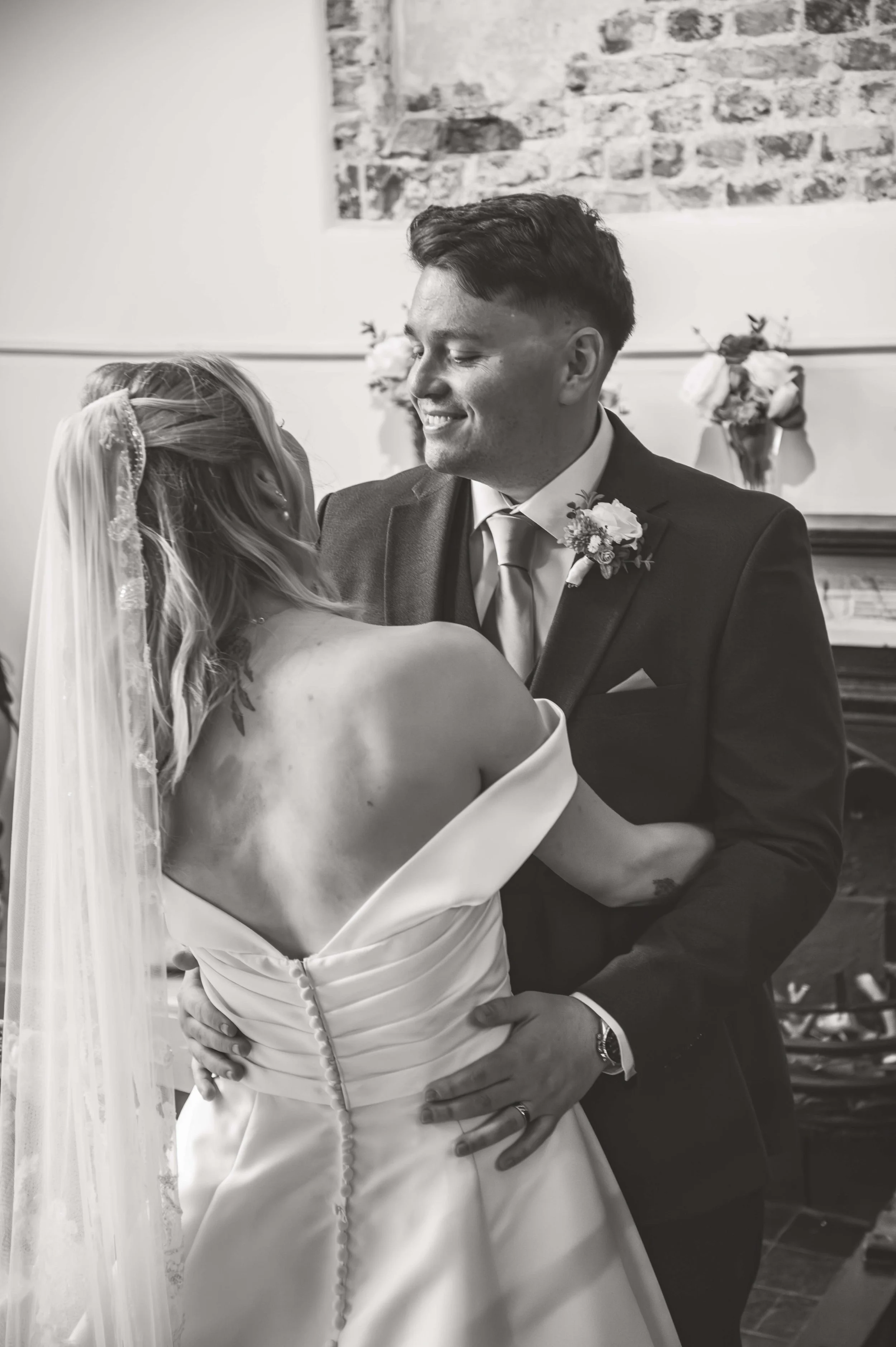 Black and white photo of a wedding couple dancing. The bride wears an off-the-shoulder wedding gown and veil, and the groom wears a dark suit with a tie and boutonniere. They are smiling and holding each other close in a romantic moment.