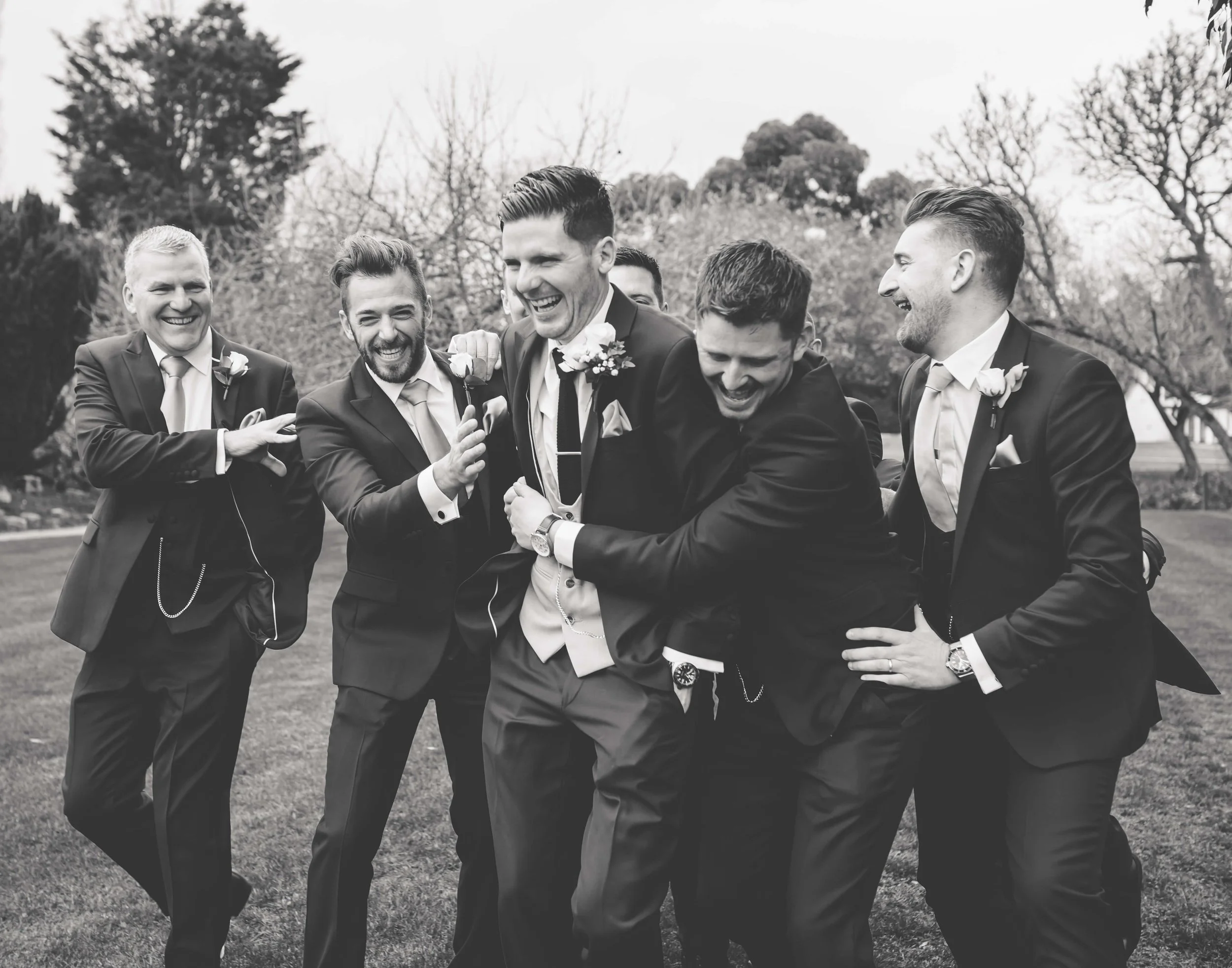 A group of grooomsmen in suits laughing and playing together outdoors on a grassy area, with trees in the background at wedding.