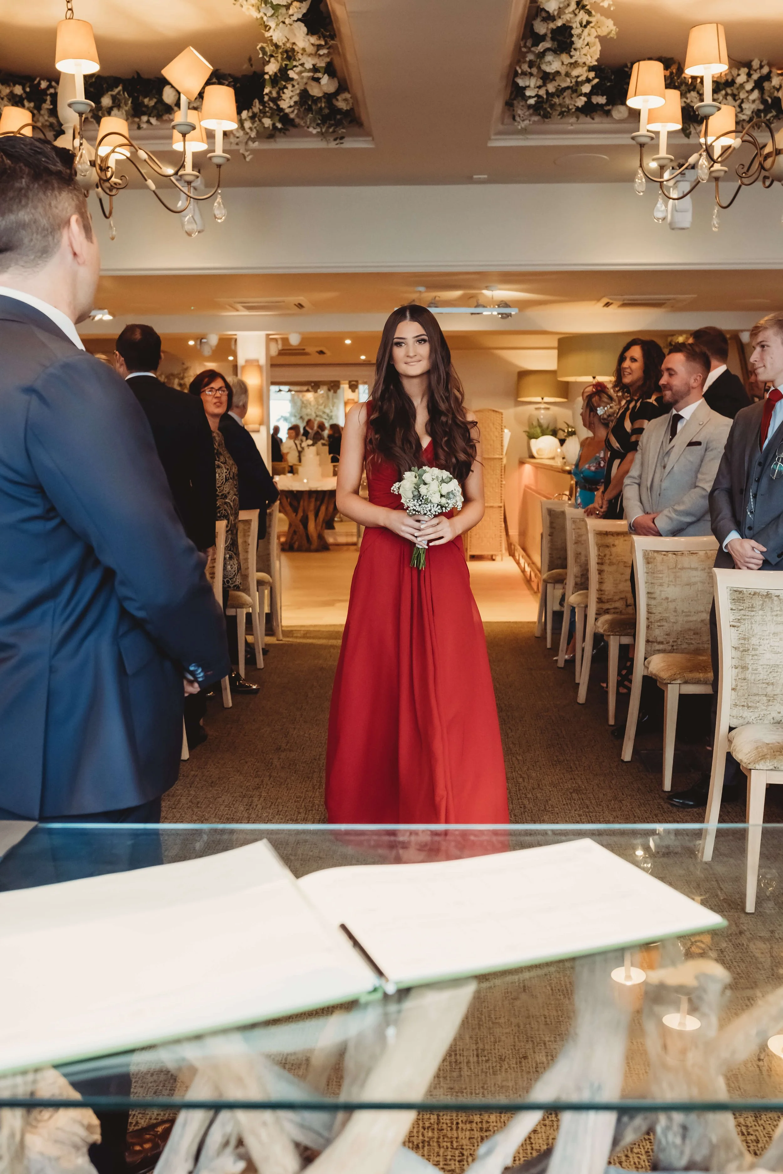 A woman in a red dress holding a bouquet of white flowers, standing in a formal setting with guests seated and standing, decorated with chandeliers and floral arrangements, during a wedding or special ceremony.