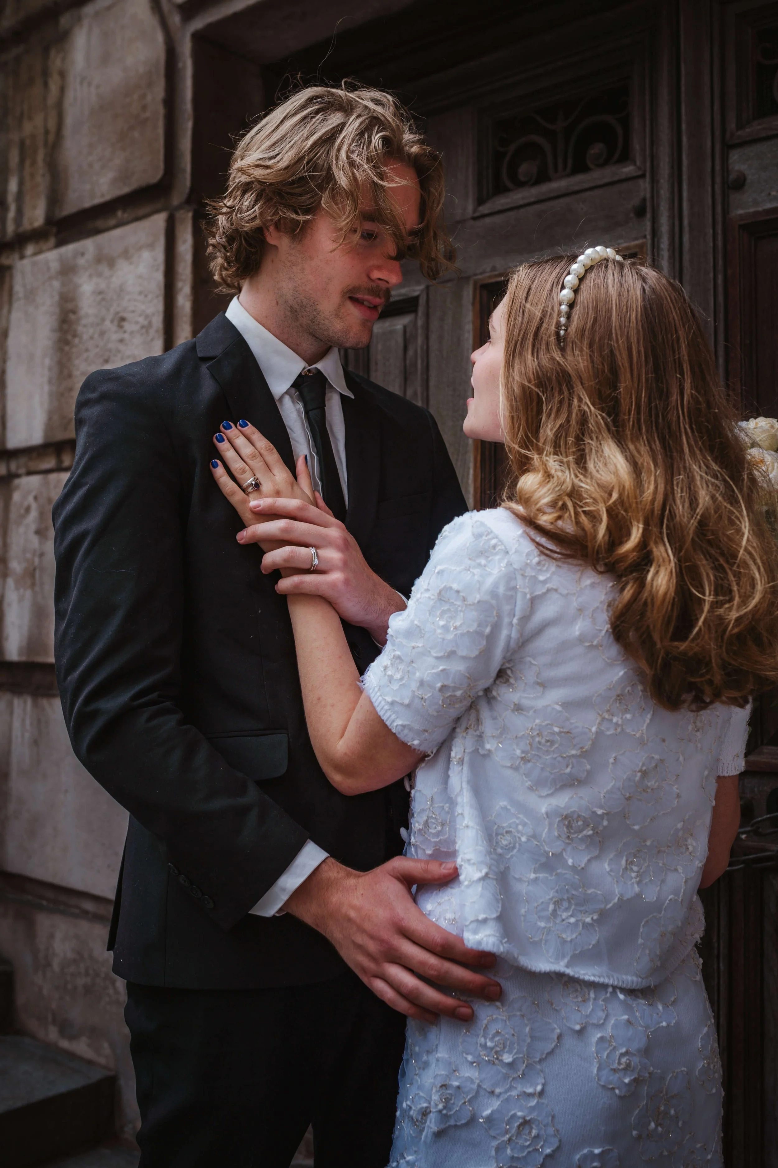 A man and woman in wedding attire are standing close to each other, looking into each other's eyes, with a stone wall and door behind them.