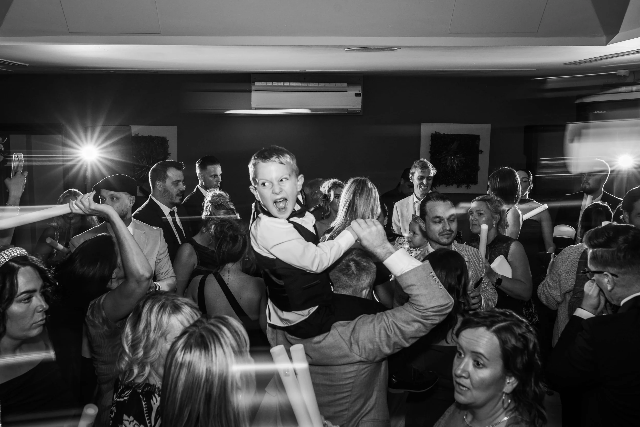 A lively party scene with many people dancing and socializing. A young boy is sitting on an adult's shoulder, excited and smiling. The gathering is indoors with bright lighting and some flash effects.