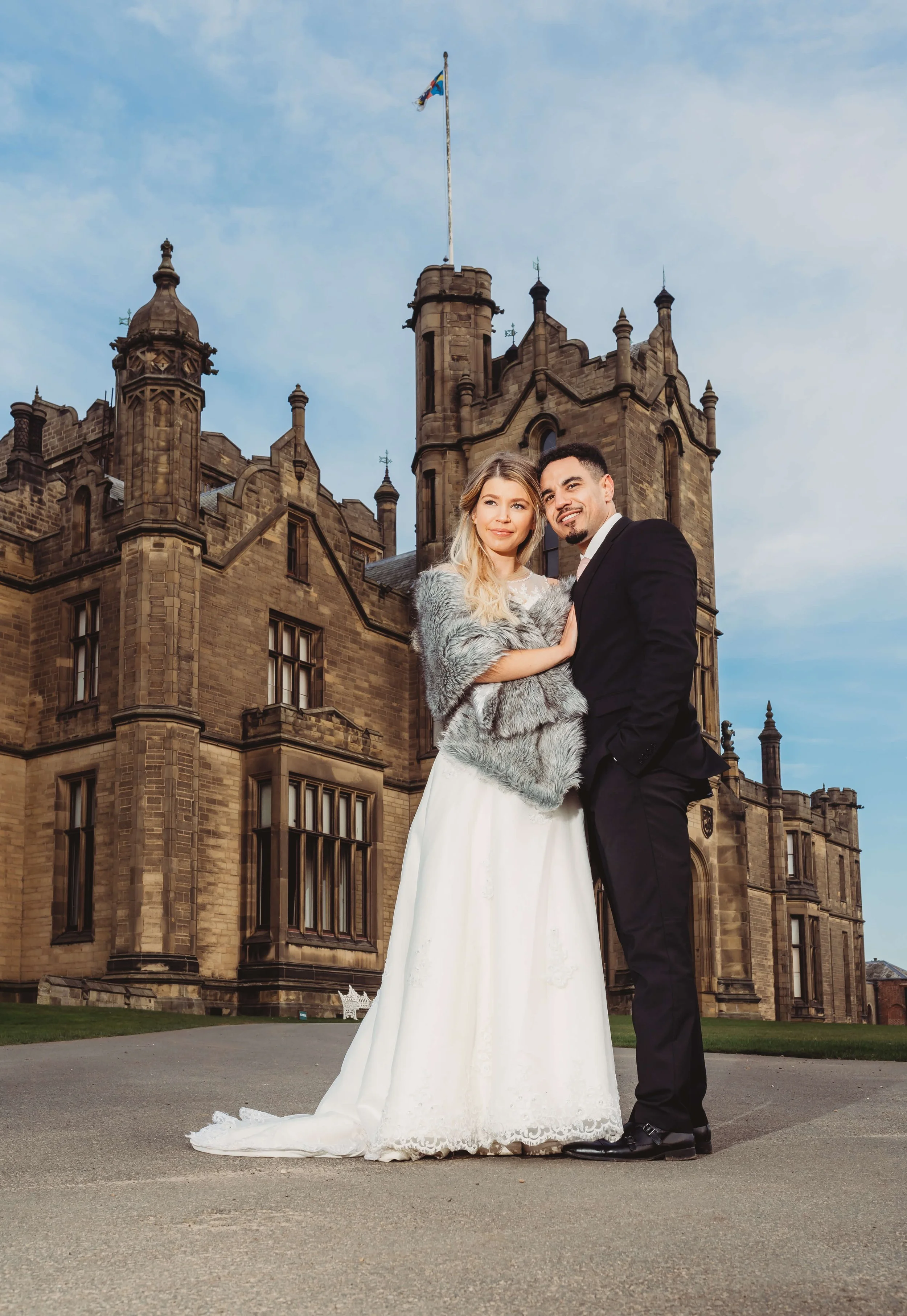 A bride and groom standing close together in front of a historic castle during sunset, with the bride wearing a white wedding dress and a gray fur stole, and the groom in a black tuxedo.