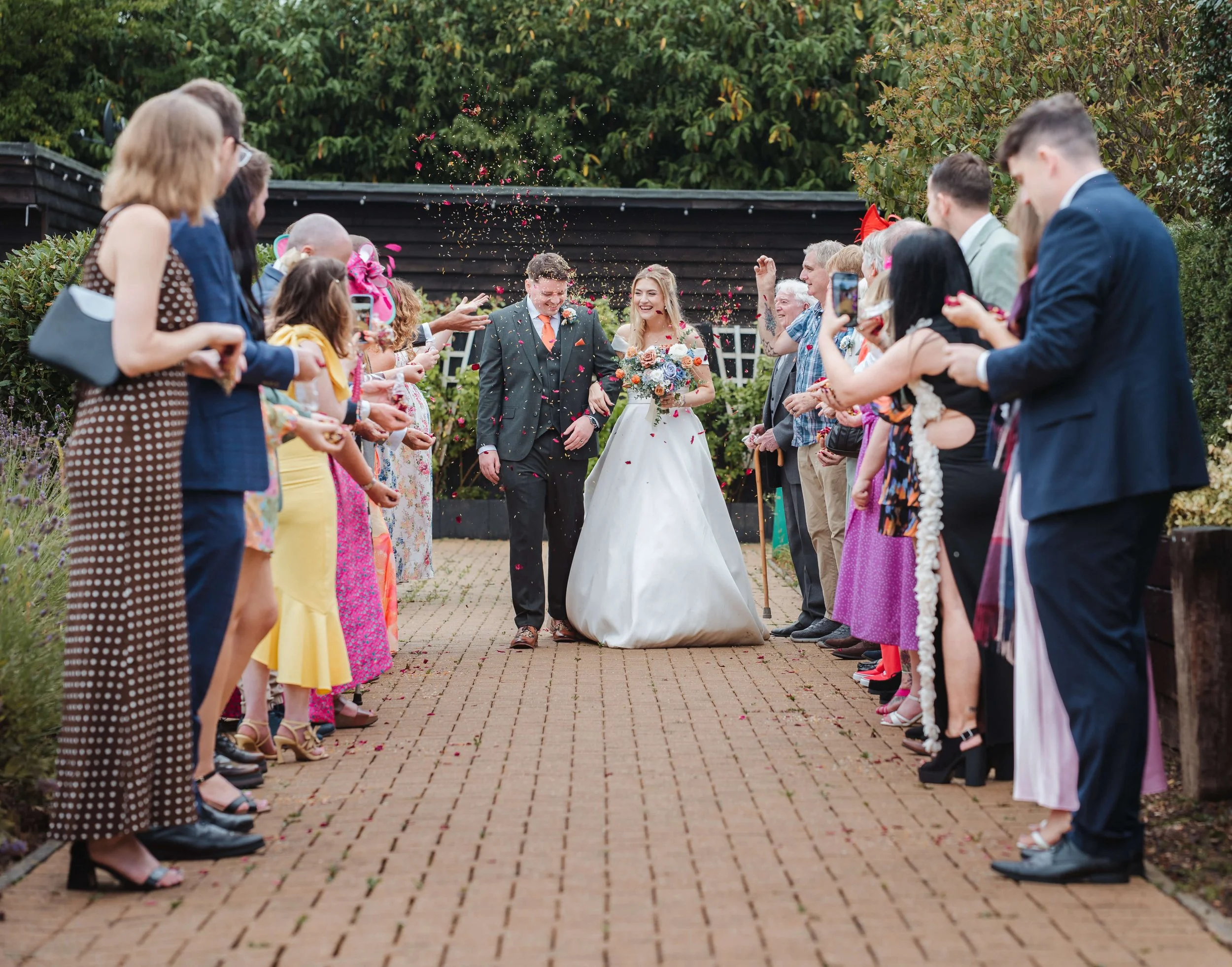 A bride and groom walking down a brick path at their wedding surrounded by friends and family throwing confetti and taking photos.