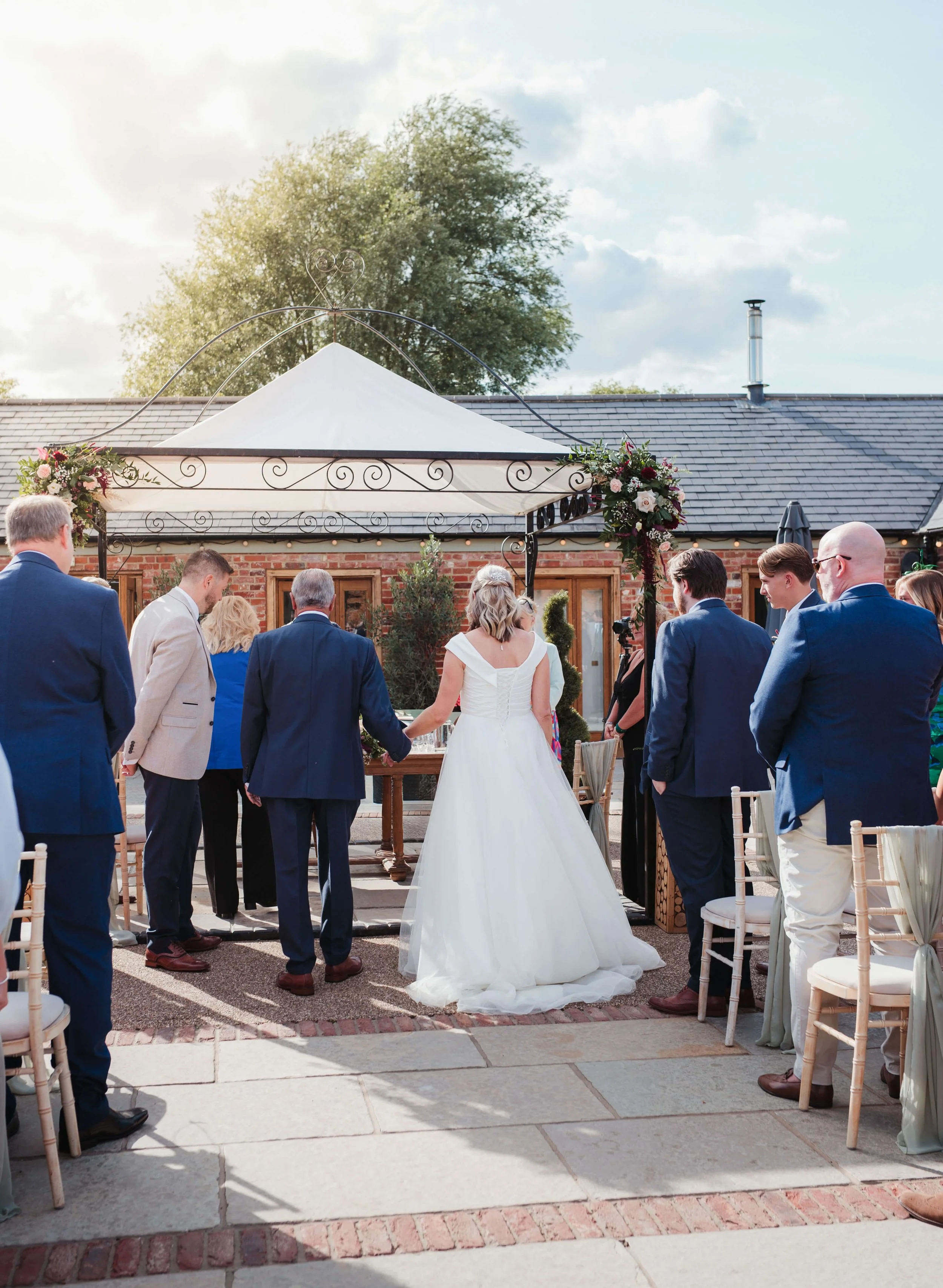 Bride and groom exchanging vows under a decorated outdoor gazebo, surrounded by guests in formal attire at a wedding ceremony.