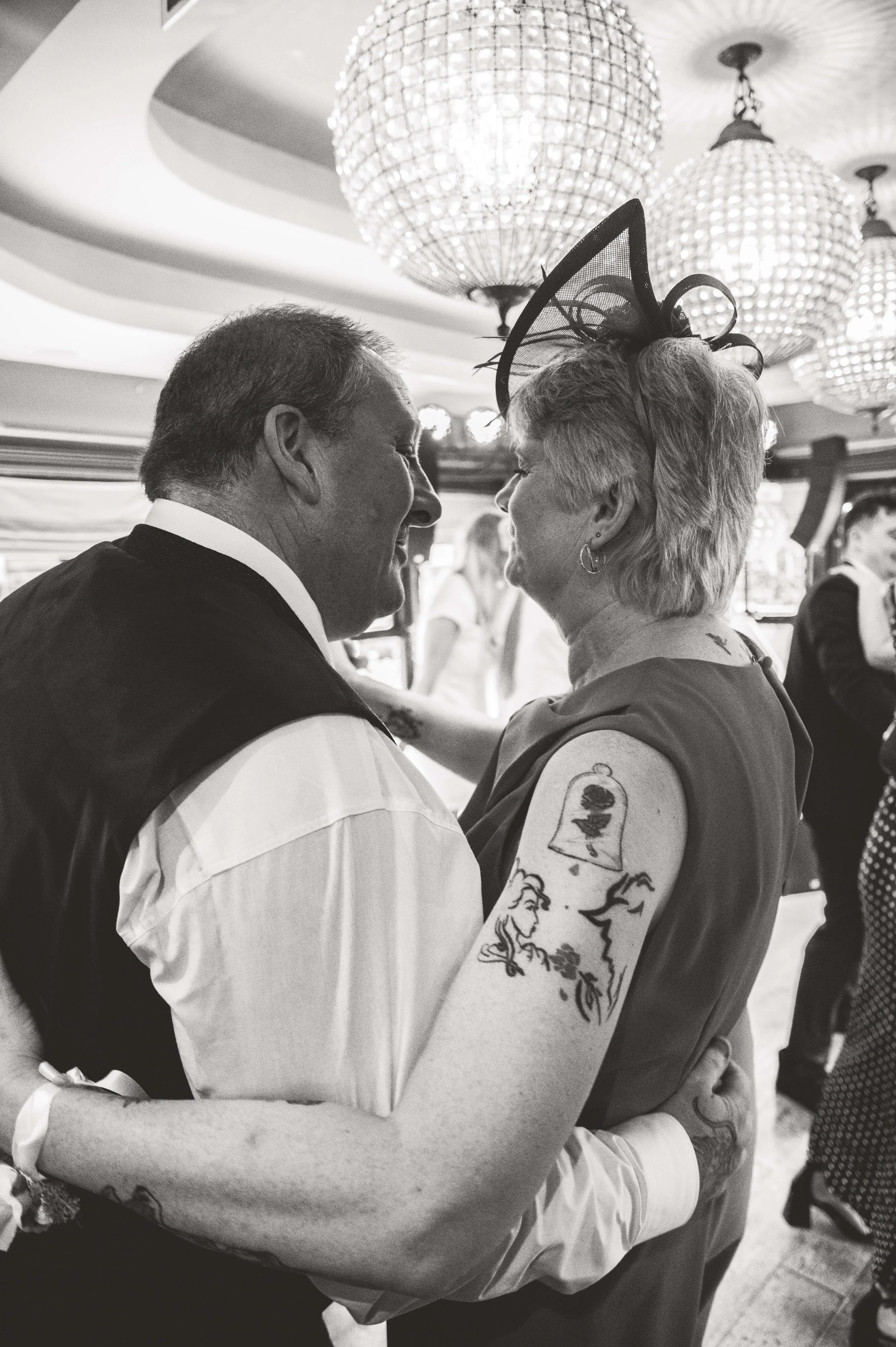 A black-and-white photo of a man and woman dancing closely, smiling at each other, in a decorated indoor setting with chandeliers, possibly at a celebration or event.