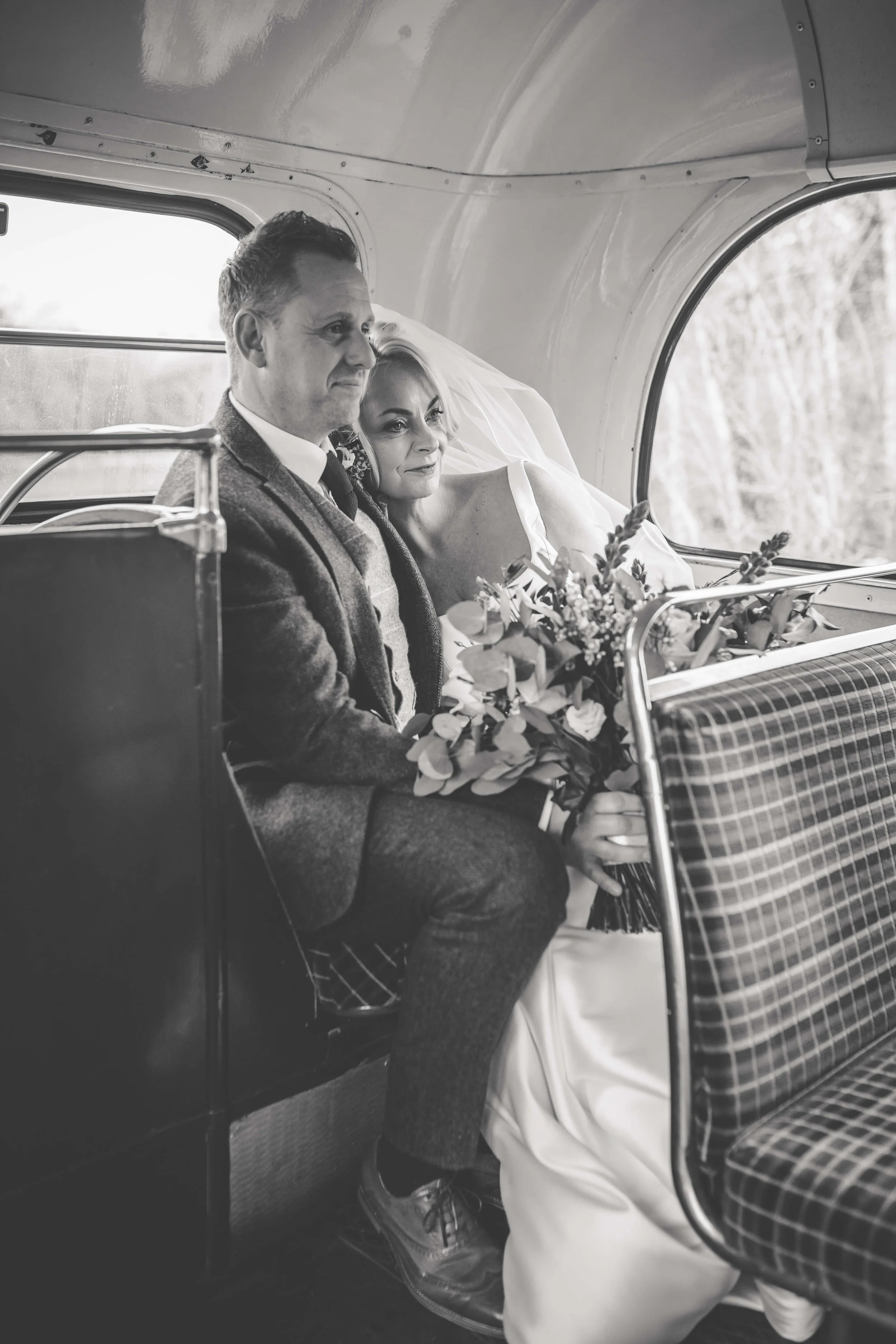 A black-and-white photo of a bride and groom sitting on a vintage train, looking out the window, with the bride holding a bouquet of flowers.