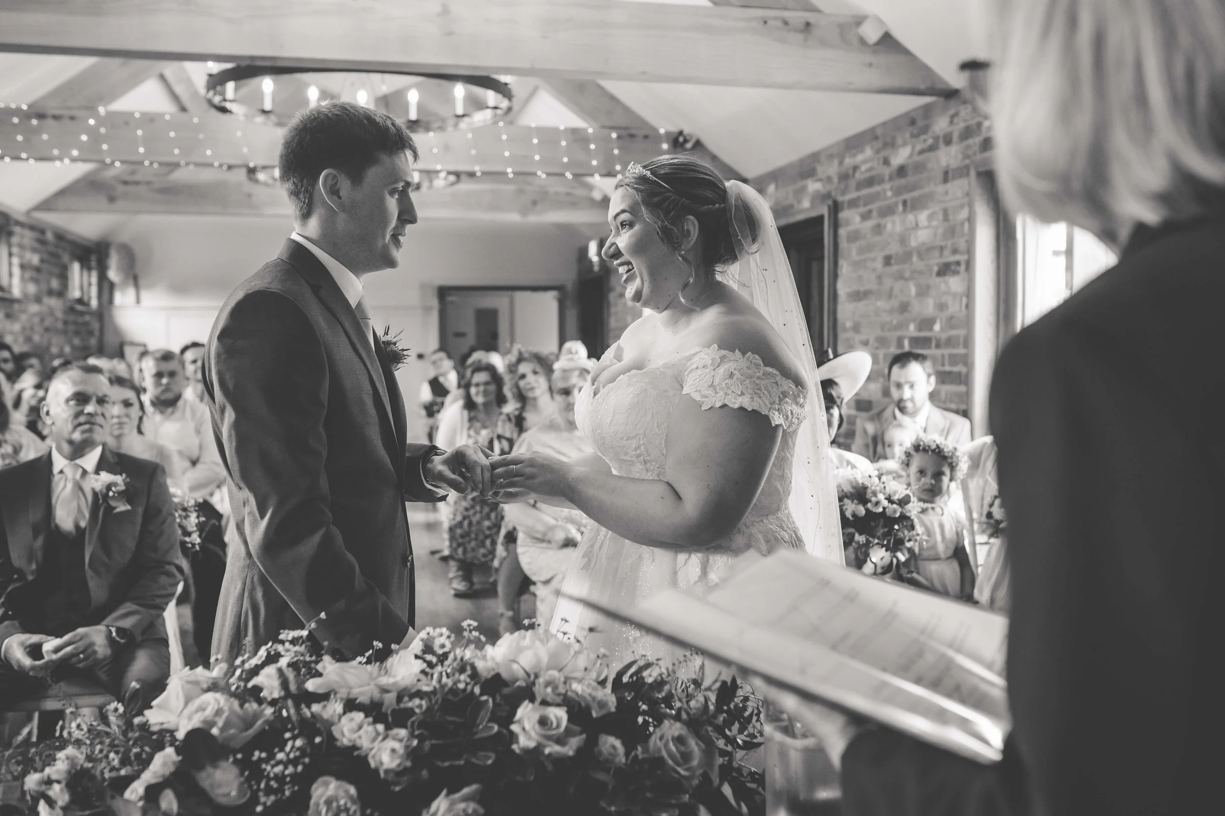 Black and white photo of a wedding ceremony with a couple holding hands and exchanging rings, surrounded by seated guests in a decorated rustic venue.
