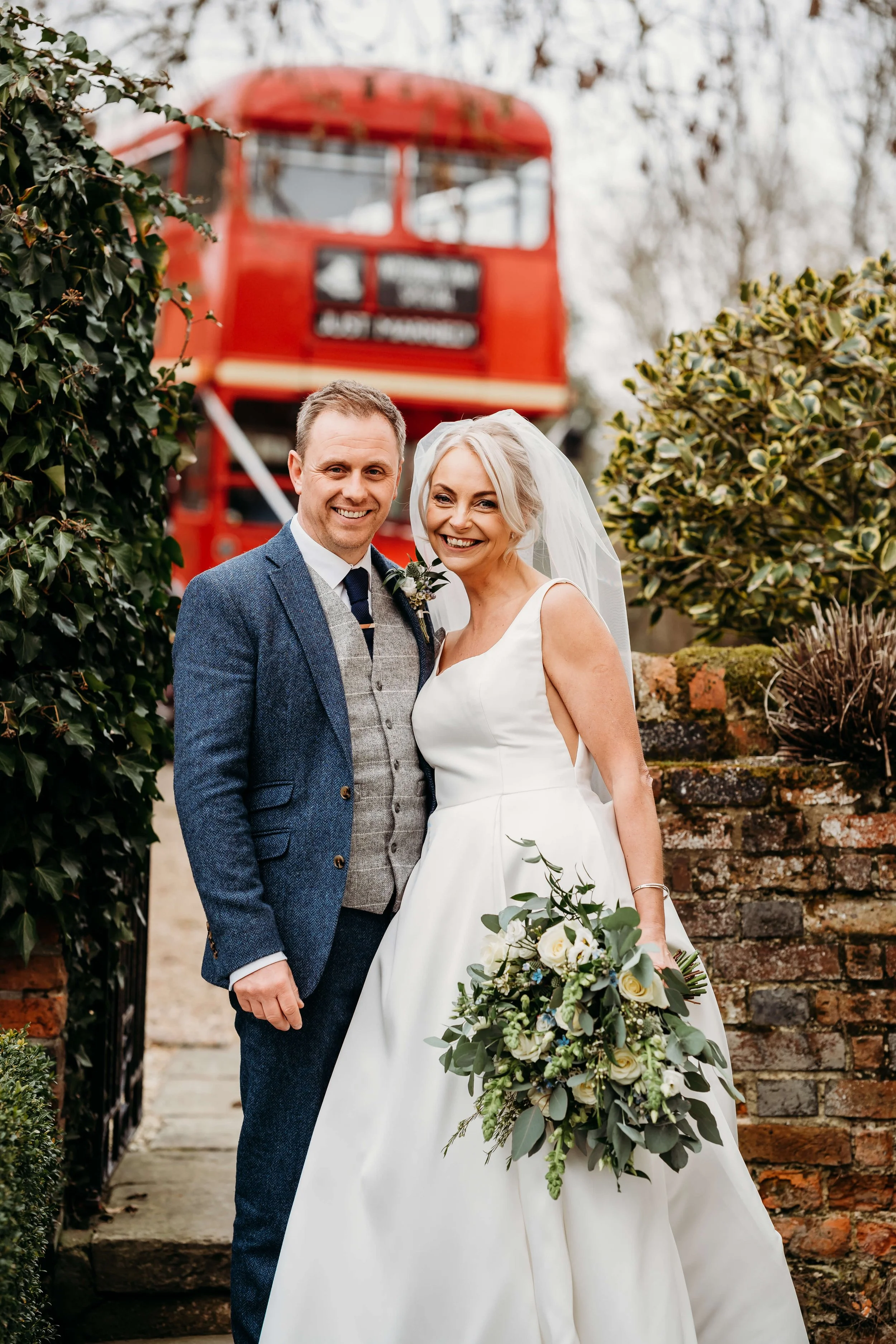 A smiling bride and groom standing outdoors, with a red double-decker bus in the background, surrounded by greenery.