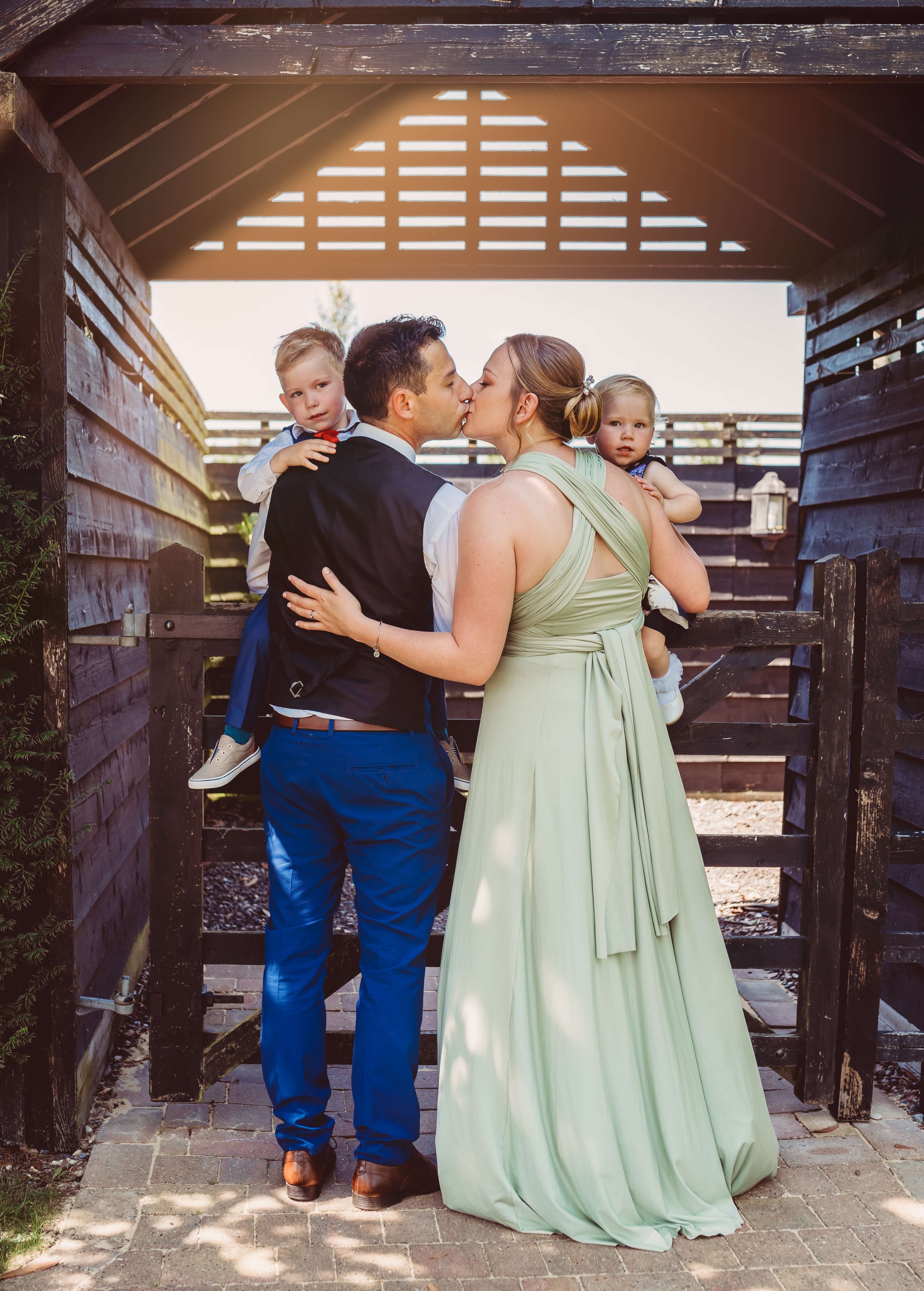 A couple sharing a kiss outdoors with two children, a boy and a girl, in a wooden enclosure.