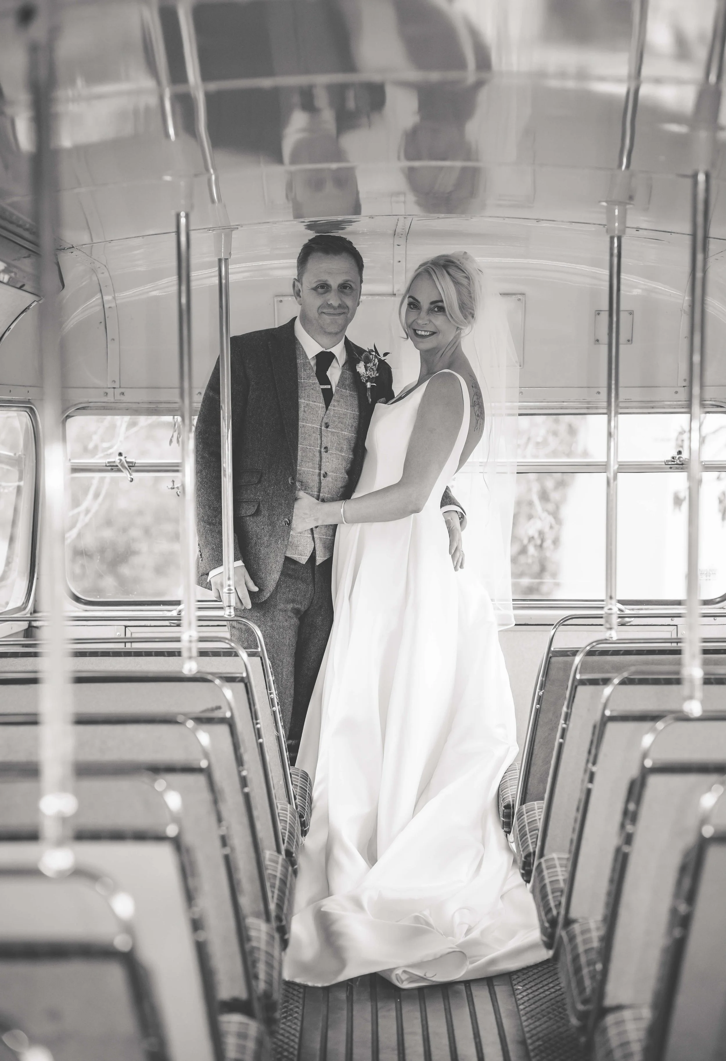 A newlywed couple on a vintage bus, with the bride in a white gown and veil, and the groom dressed in a suit with a vest and boutonniere. They are smiling and standing close together, framed by the bus's interior and windows.