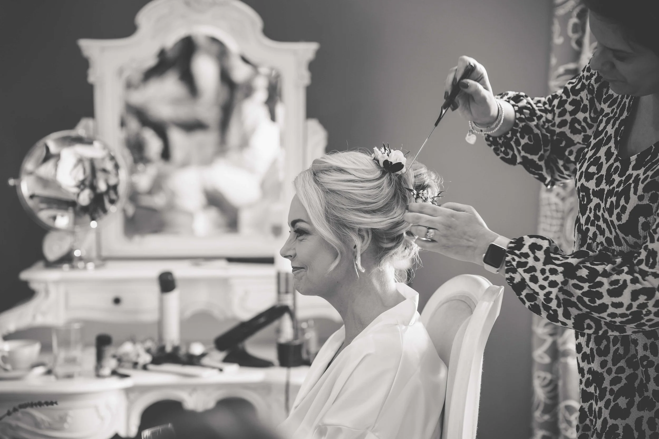 A woman getting her hair styled with flowers by a stylist in a room with a vanity mirror and makeup items.