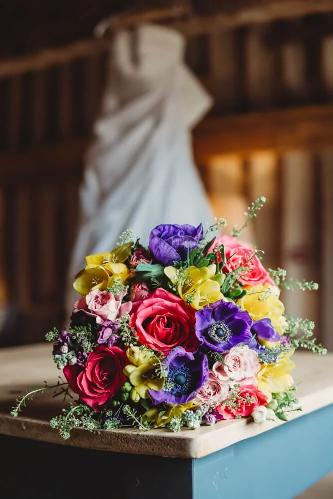 Colorful bouquet of pink, purple, yellow, and white flowers with greenery on a wooden table, with a blurred background of a rustic barn interior and a white hanging draped fabric.