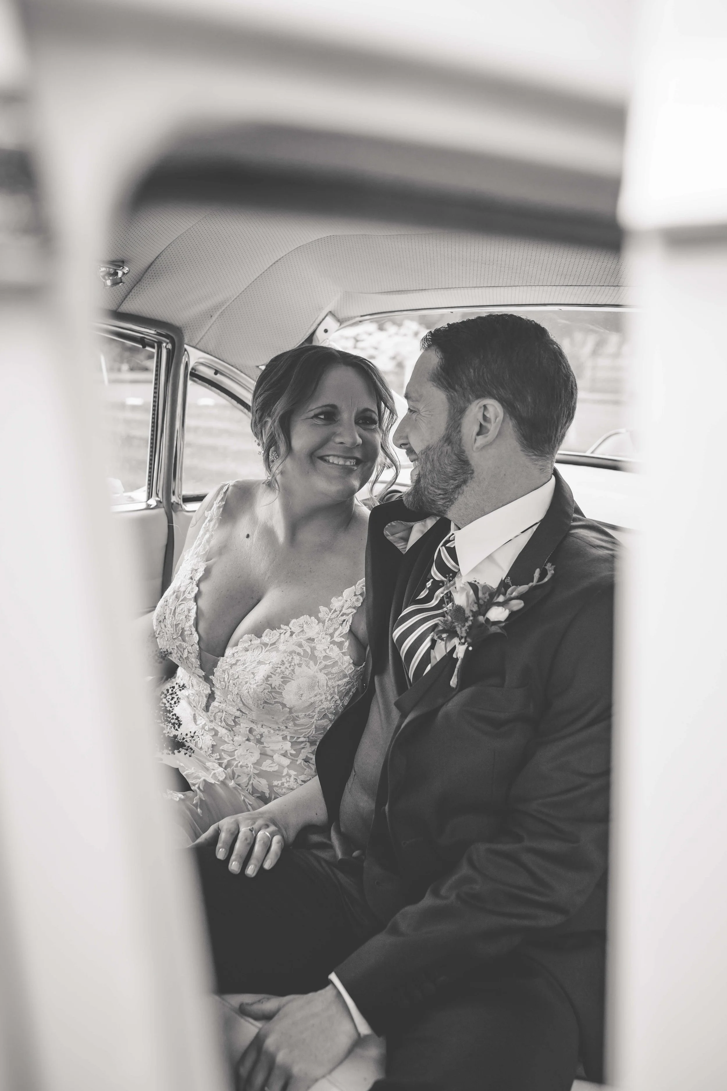 Black and white photo of a smiling bride and groom sitting close together inside a car, looking at each other lovingly, as seen through the car window.