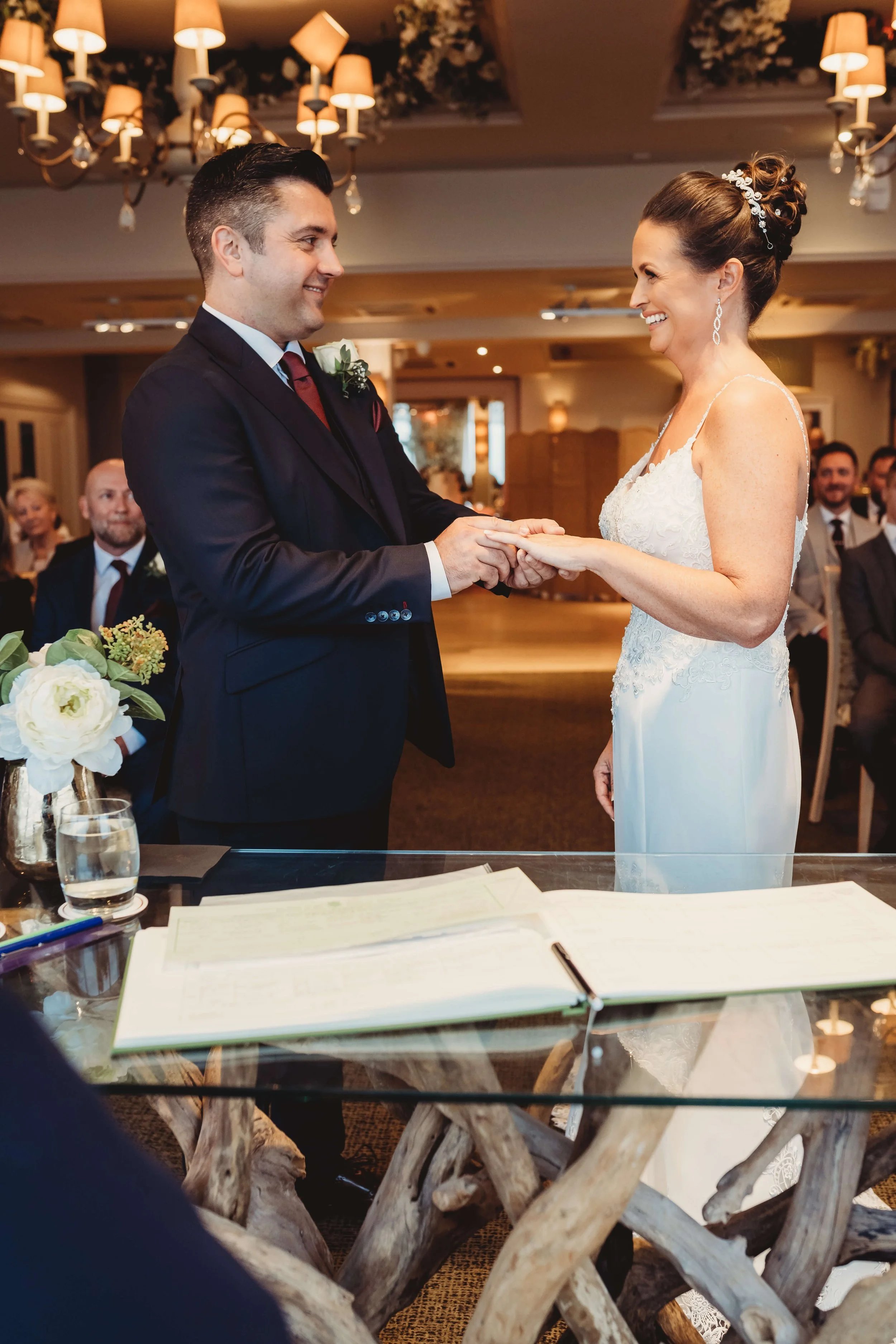 A wedding ceremony with a groom and bride exchanging rings, surrounded by seated guests in a decorated indoor venue.