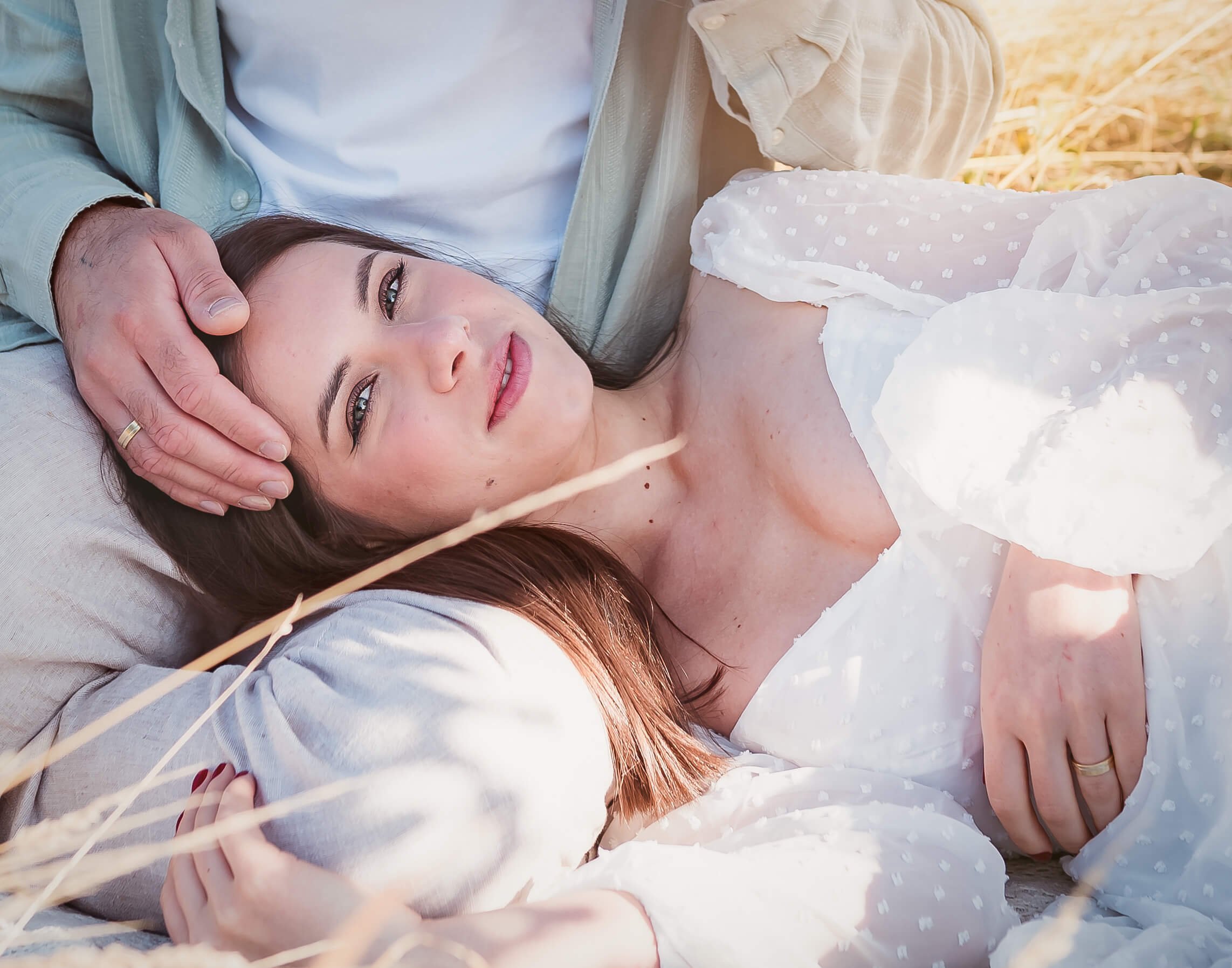 A woman lying on the ground outdoors, gazing into the camera, with a man resting his hand gently on her forehead.