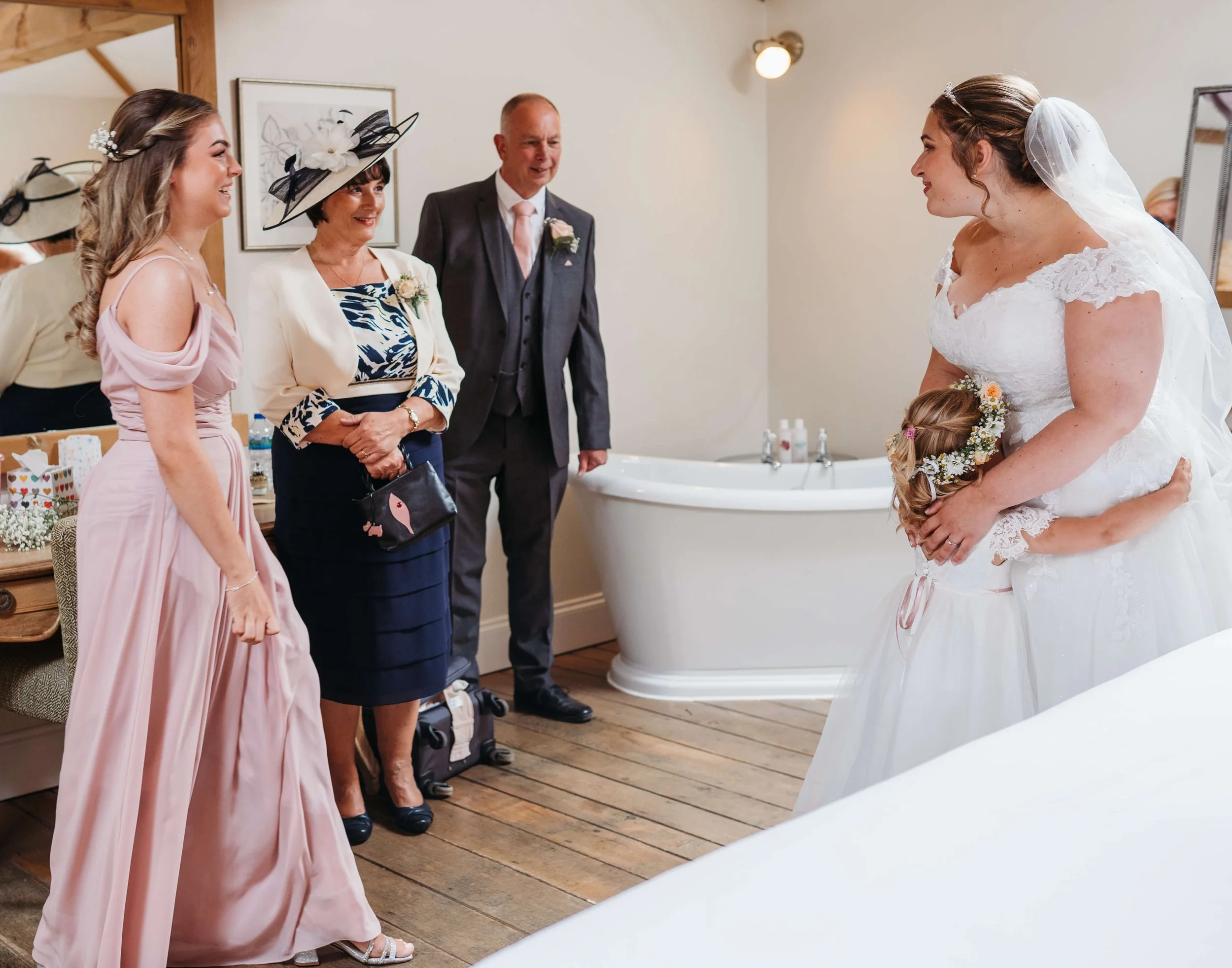 Bride and a young girl, possibly her daughter, in wedding dresses, standing together and facing each other in a bedroom with a bathtub in the background. Four other people, two women and two men, are standing nearby, dressed in formal attire, observi