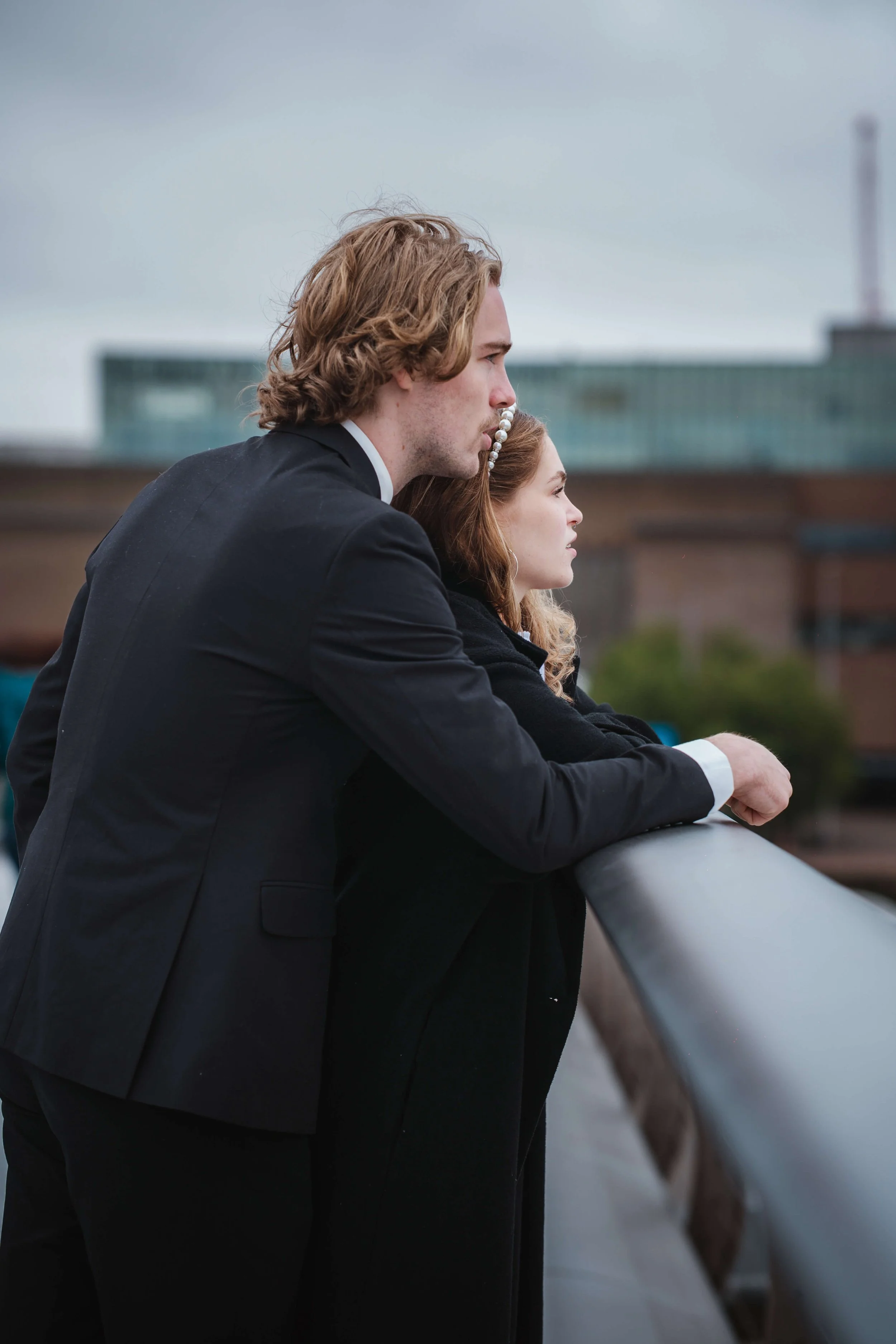 A young man and woman stand side by side on a bridge, looking out over the city with a serious expression on their faces. The man is dressed in a black suit, and the woman has red hair with a pearl headband.