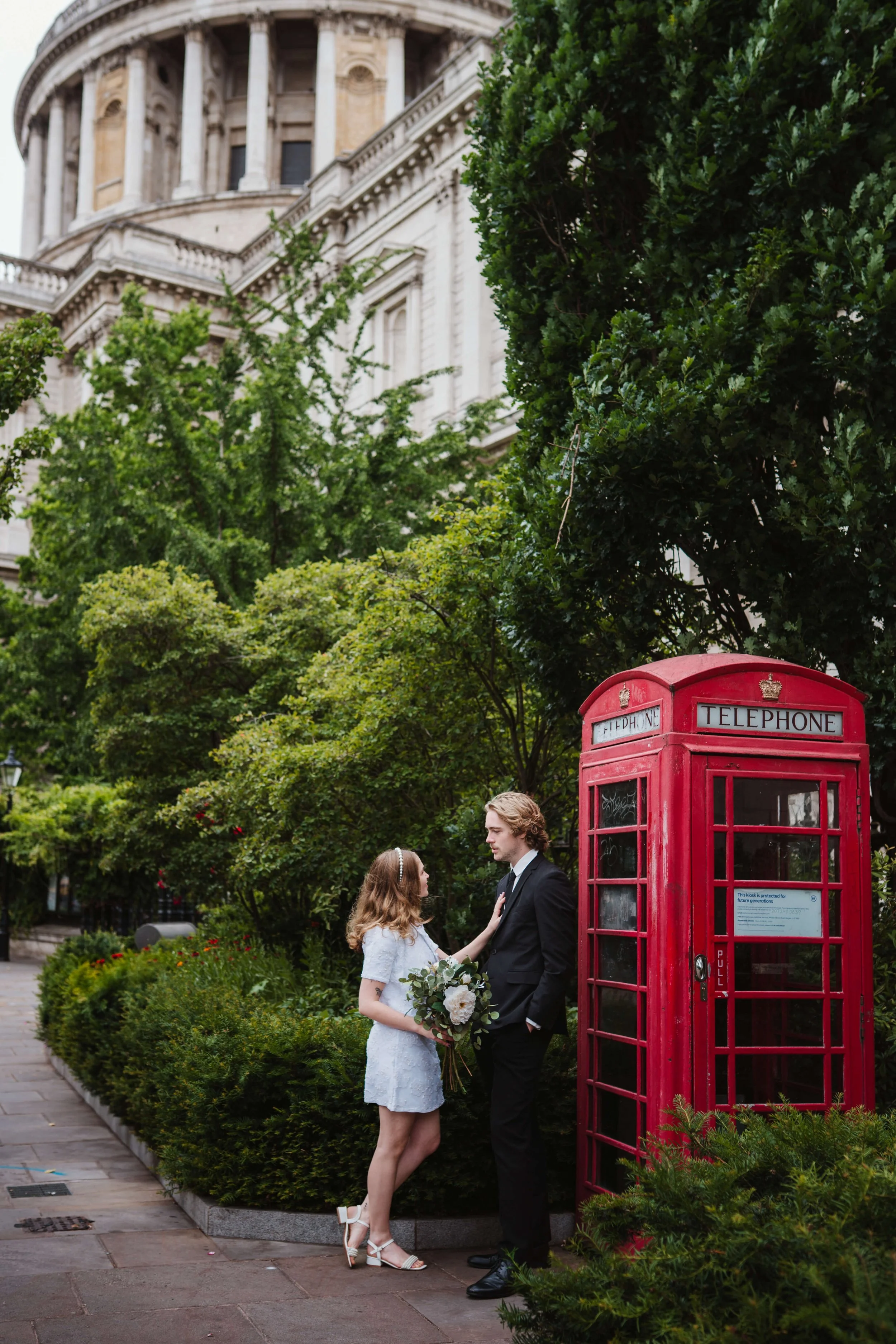 A couple dressed in formal attire standing outside near a red telephone booth, with the woman holding a bouquet of white flowers and the man wearing a black suit, in a lush, green park feeling romantic.