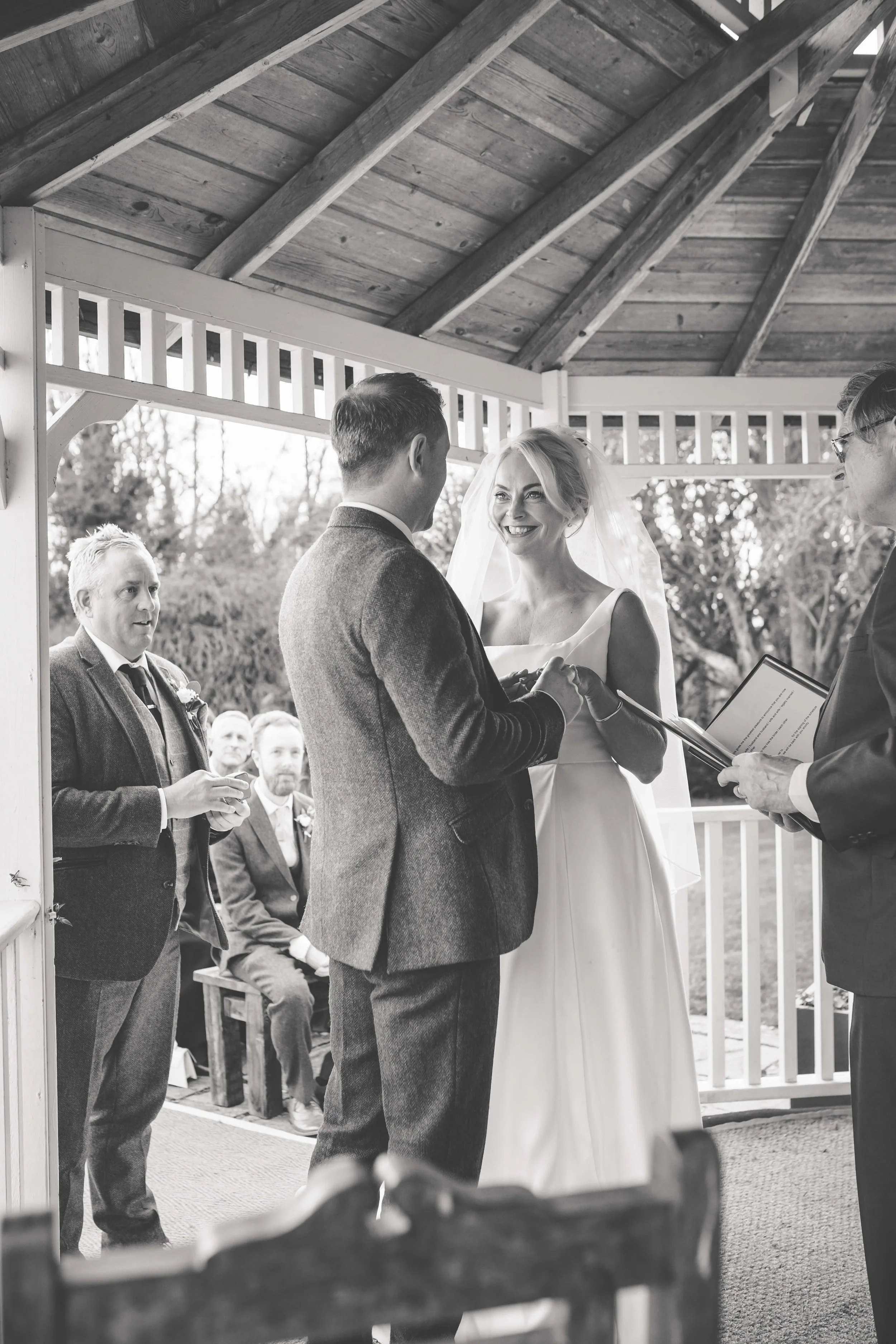 A black and white photo of a wedding ceremony outdoors, showing a bride and groom exchanging vows under a wooden pavilion, with officiant holding a book, and guests seated in the background.