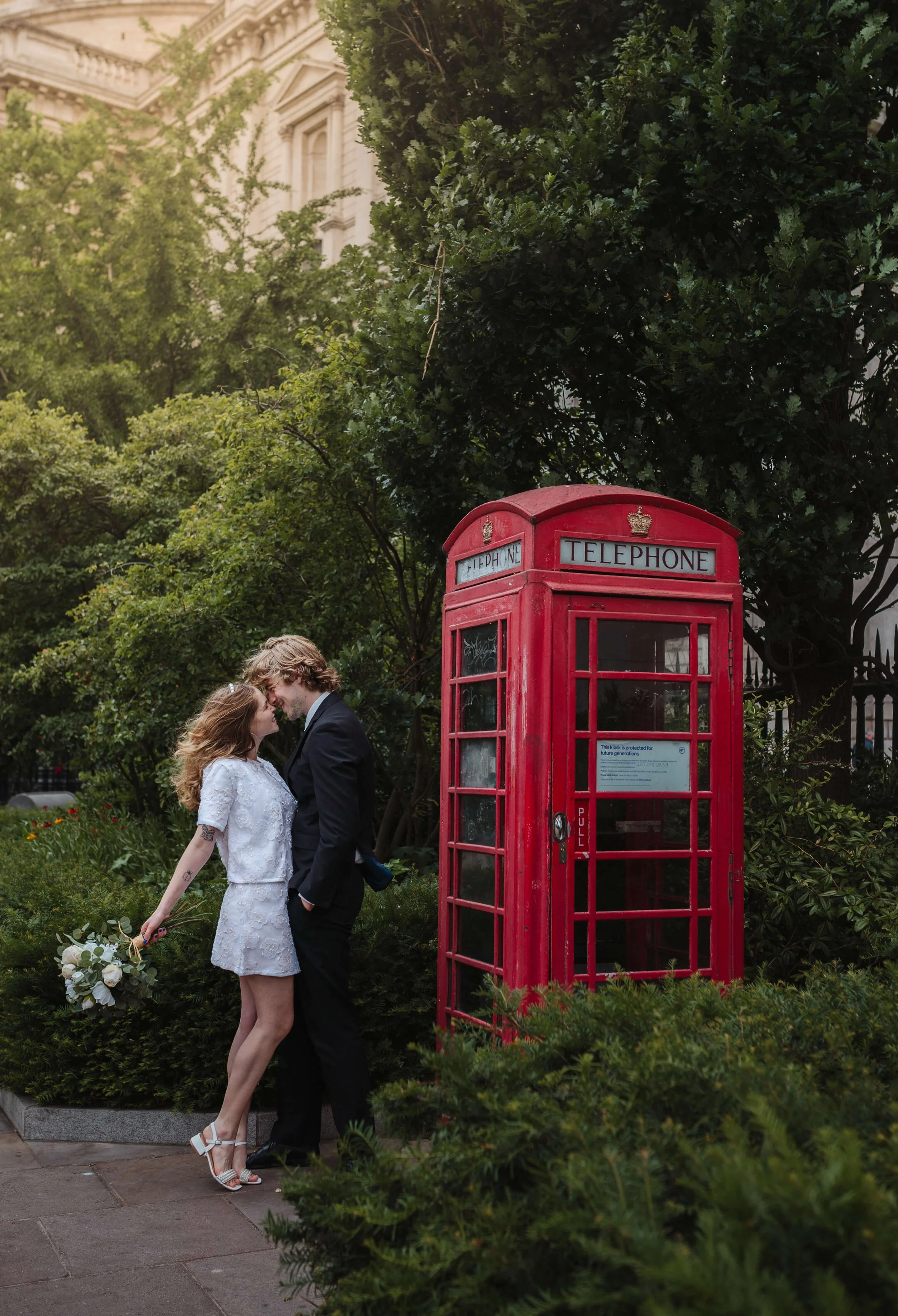 A couple with wedding attire standing close and touching foreheads beside a red telephone booth in a garden.